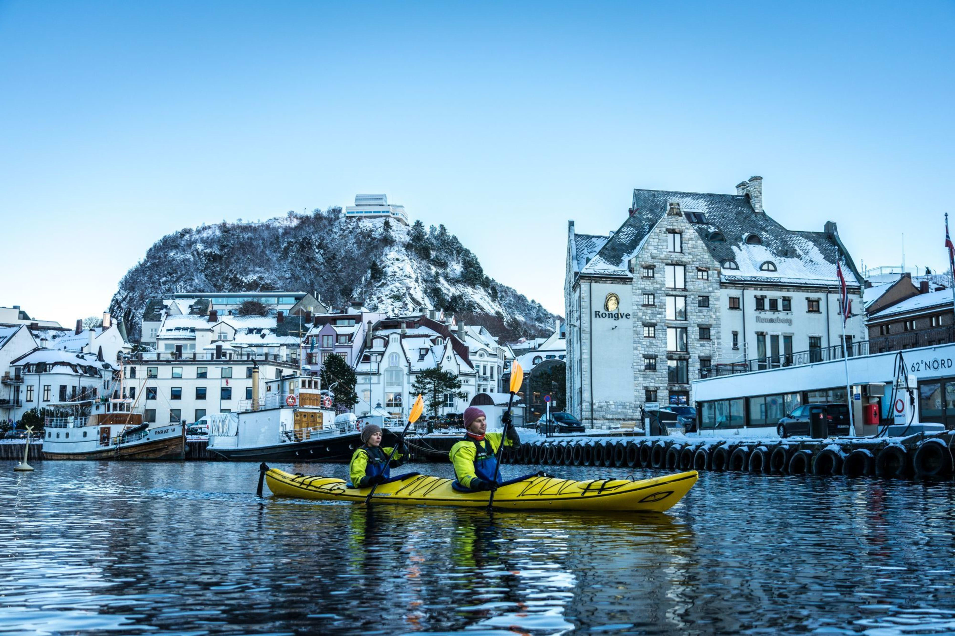 Two people paddle a yellow tandem kayak through calm waters, with snow-covered buildings and boats around them.