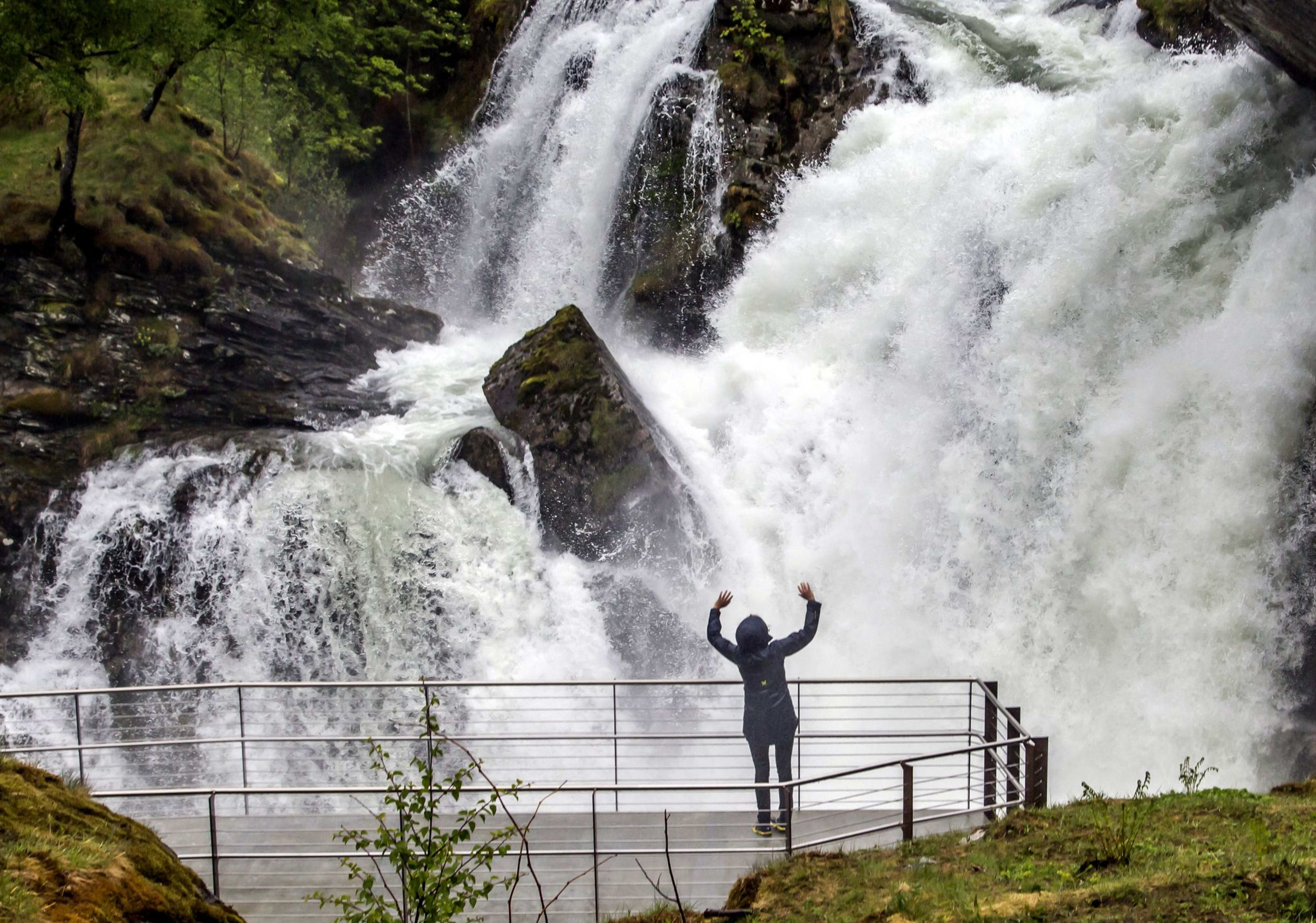 Person in Regenjacke hebt die Arme vor gewaltigem Wasserfall im Grünen.