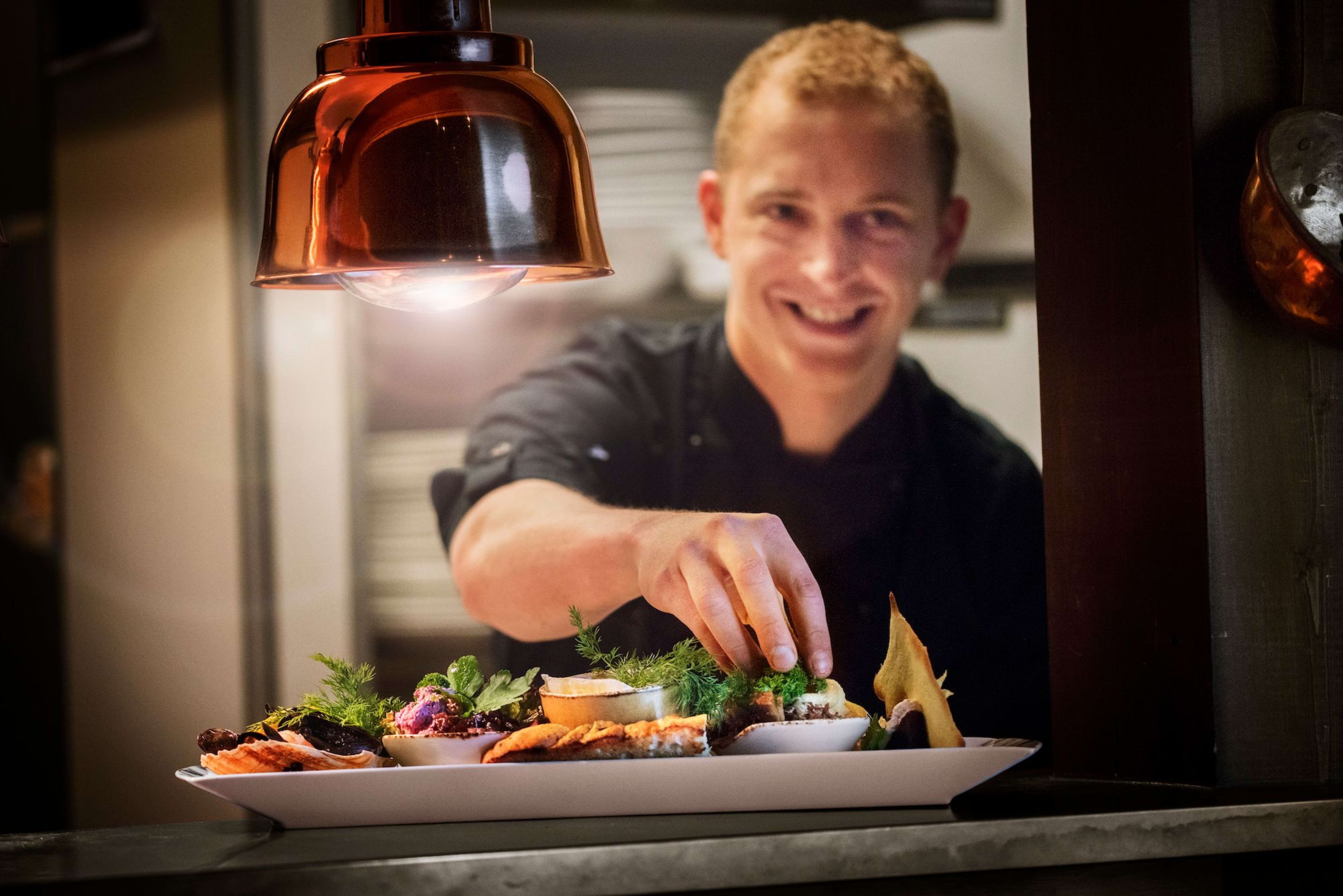 Kokk presenterer gourmetfat under varm lampekonstruksjon, fylt med delikatesser og friske urter, i koselig kjøkken.