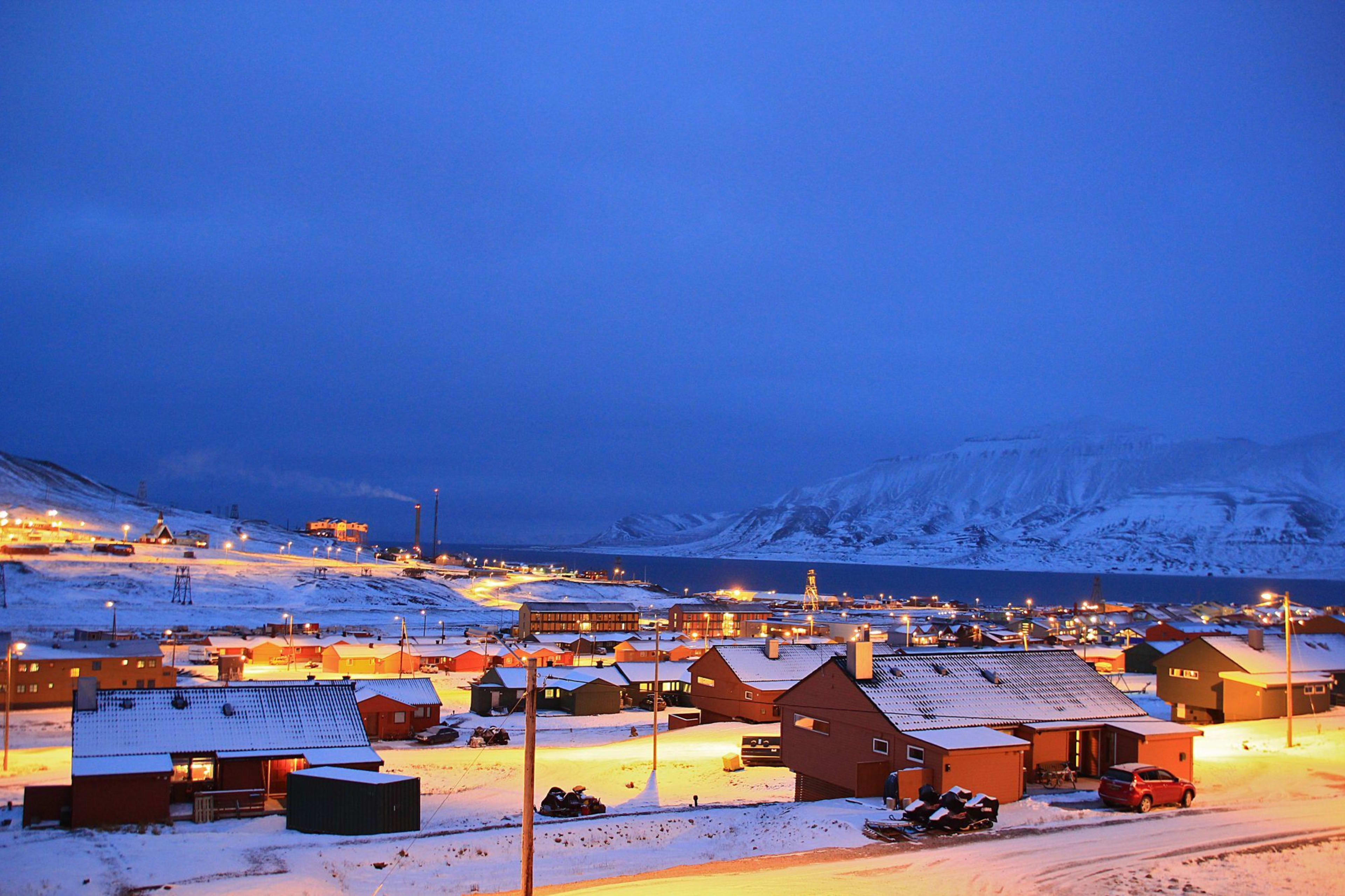 Lite by dekket med snø om natten, varmbelyste hus, gatebelysning som kaster et mykt lys, med snødekte fjell i bakgrunnen.