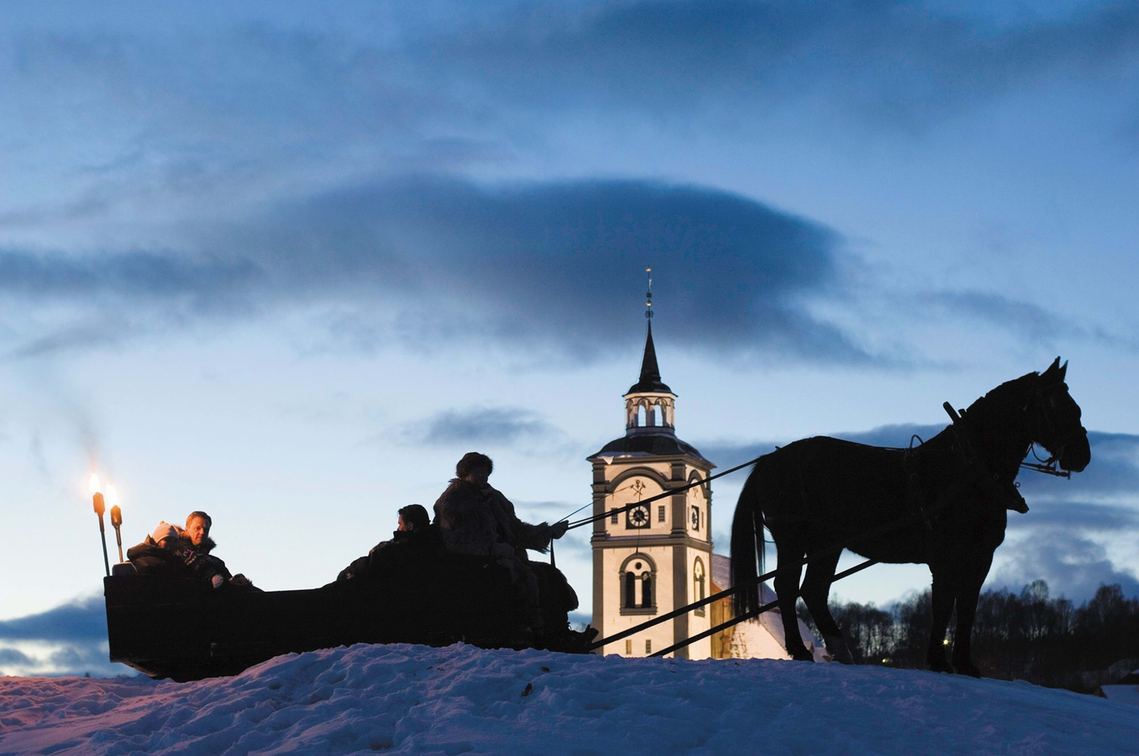 People riding in horse-drawn sleigh through snow, with torches lit, church spire, and peaceful winter landscape.