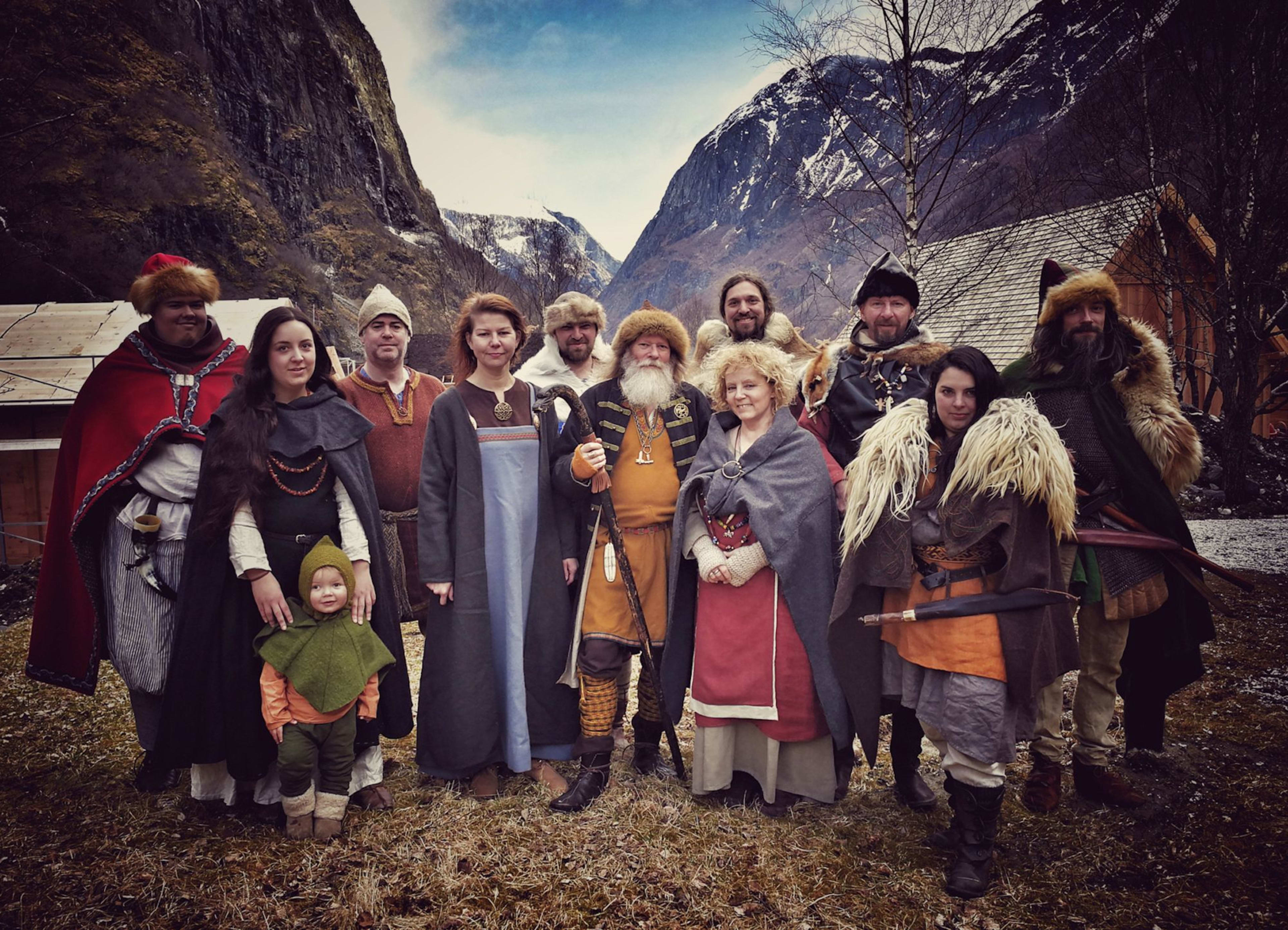 Group dressed in Viking-style clothing, posing outdoors with snow-dusted mountains, cabins, and cloudy sky in the background.