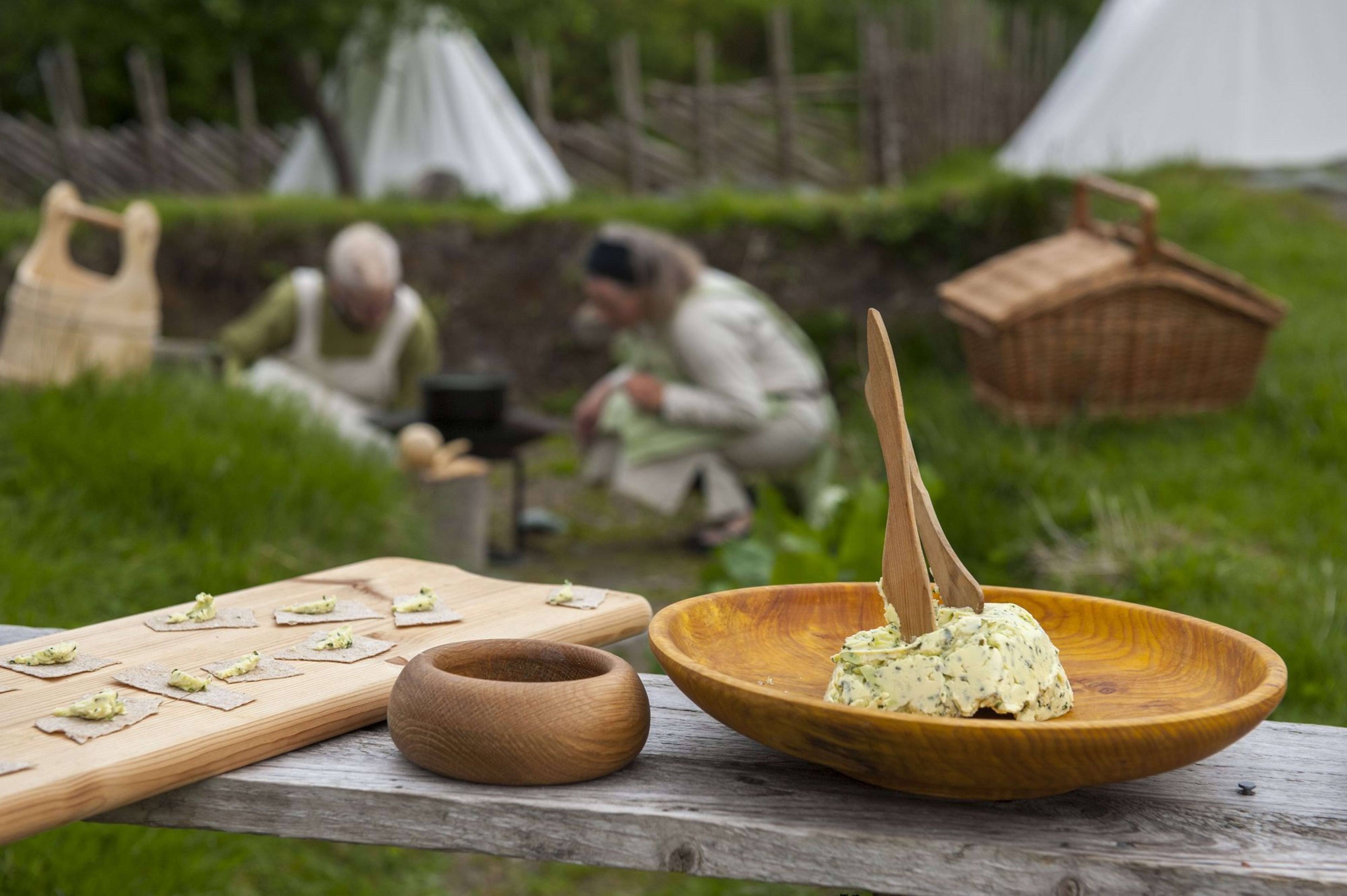 Tre Vikingbord med matforberedelse, urtesmør, treboller og kjeks, med historiske figurer i bakgrunnen.