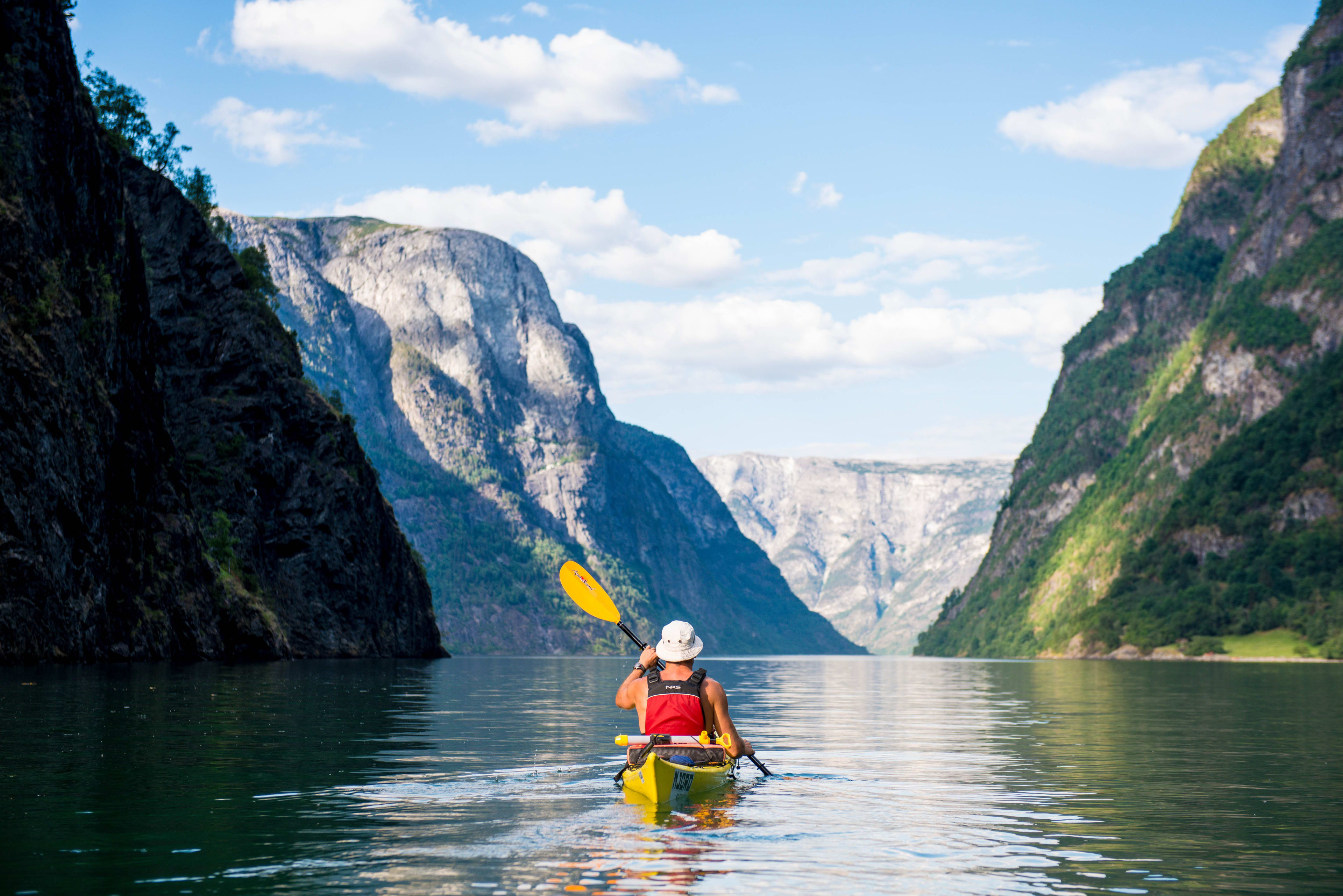 Kayaker in yellow kayak paddling through clear waters, surrounded by cliffs with greenery and mountains under blue sky.