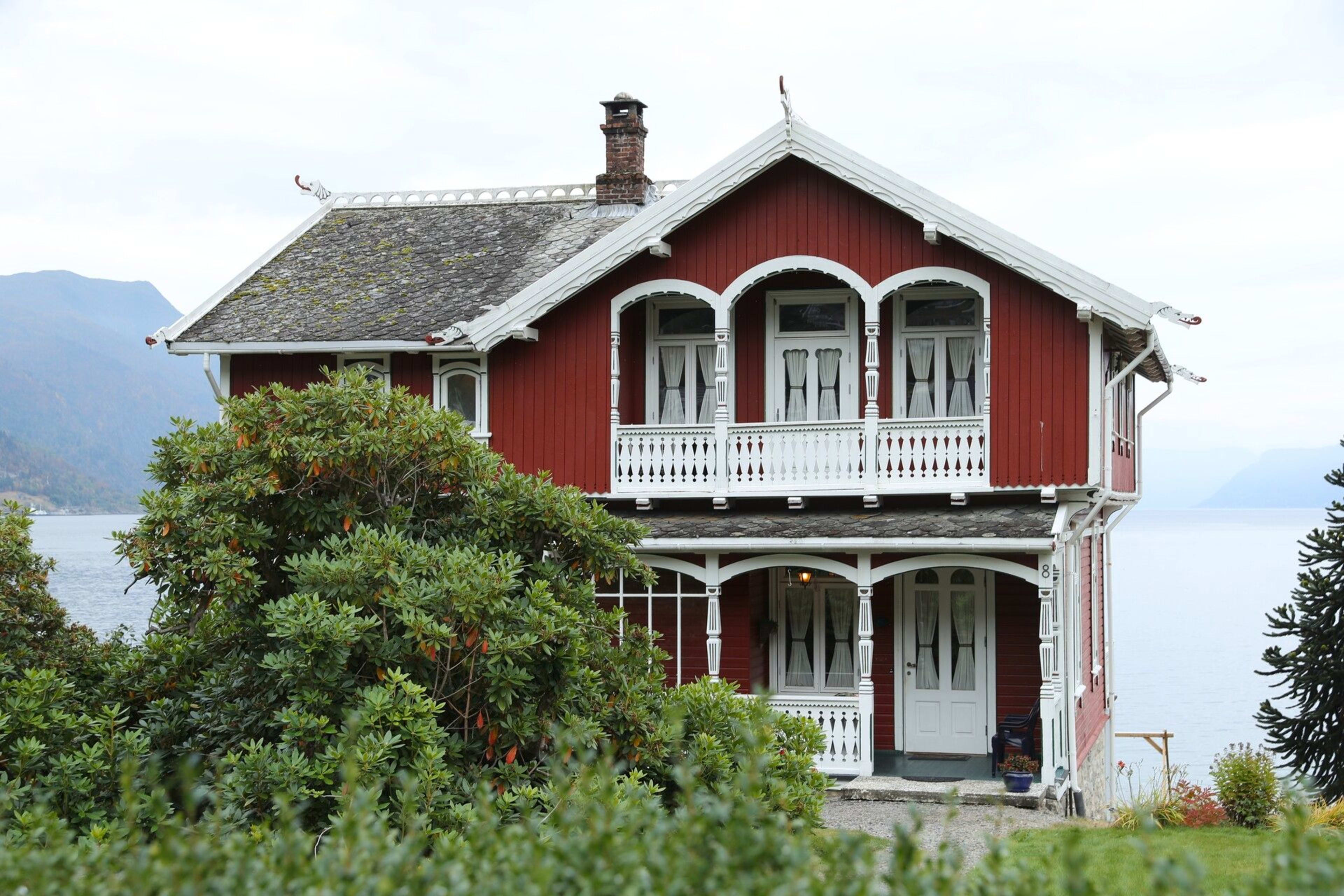 Red wooden house with white trim by fjord and mountain view.