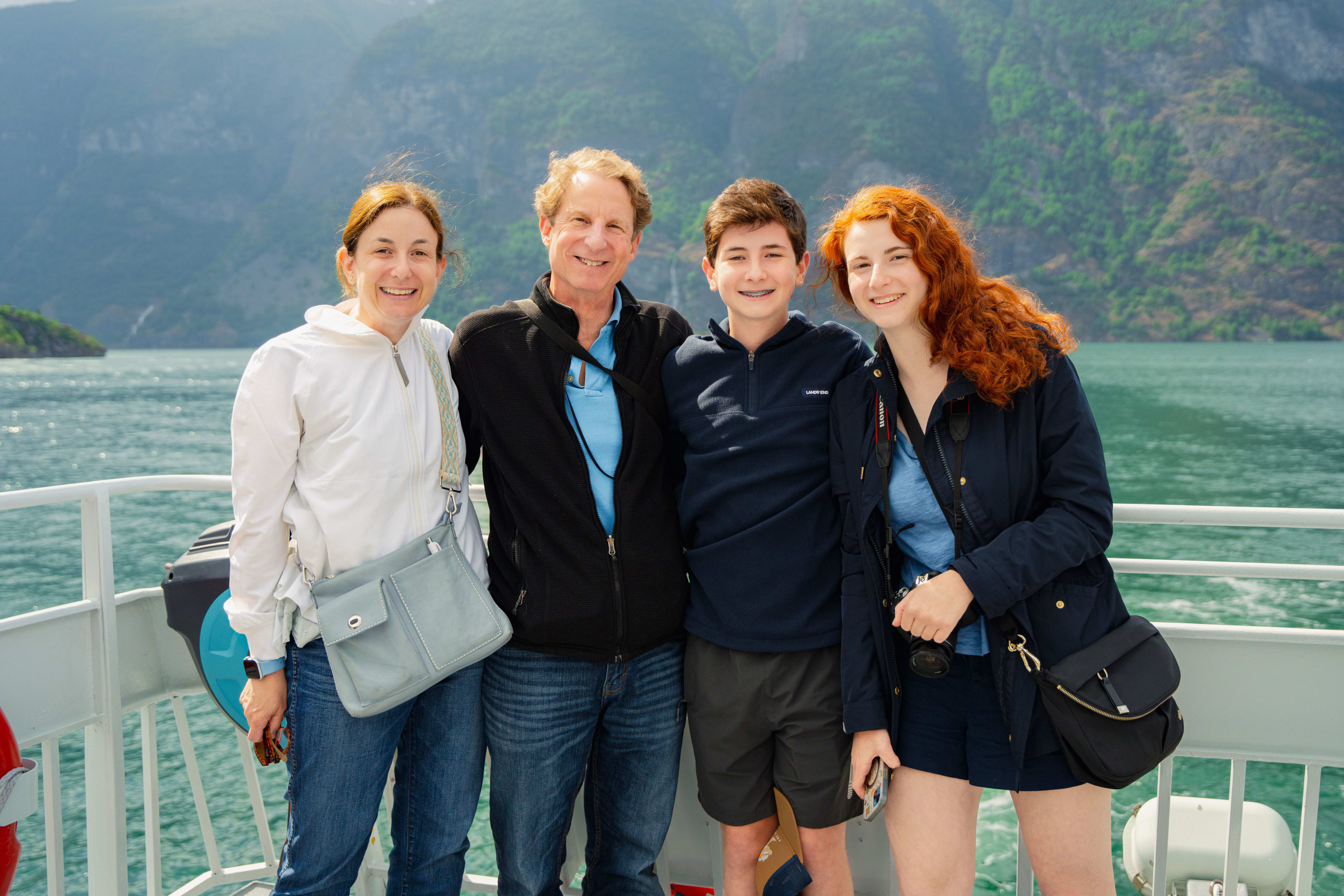 Smiling family poses on boat deck with green mountains and blue sky.