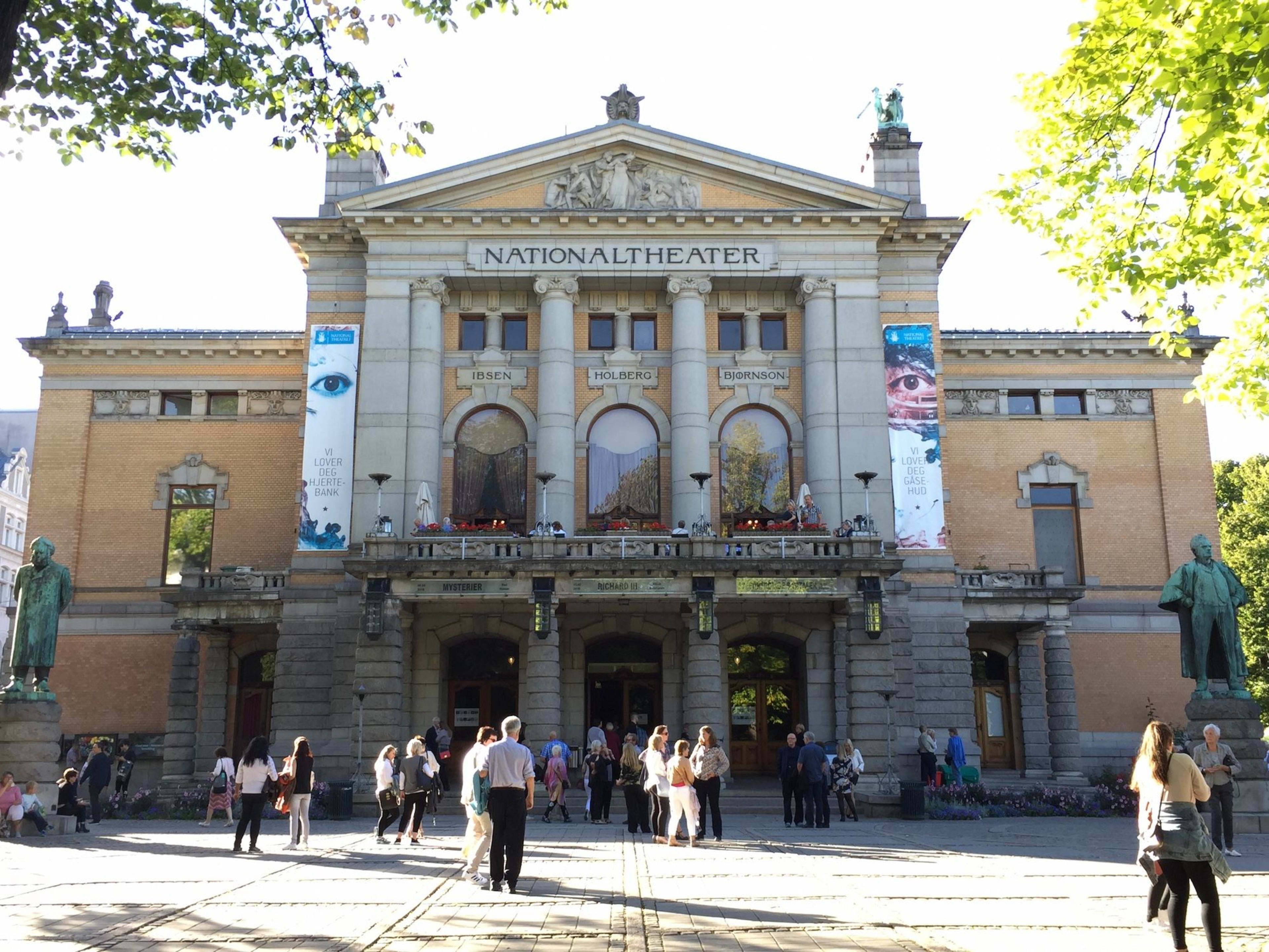Nationaltheatret i Oslo med folk utenfor på en solrik dag og statuer ved inngangen.