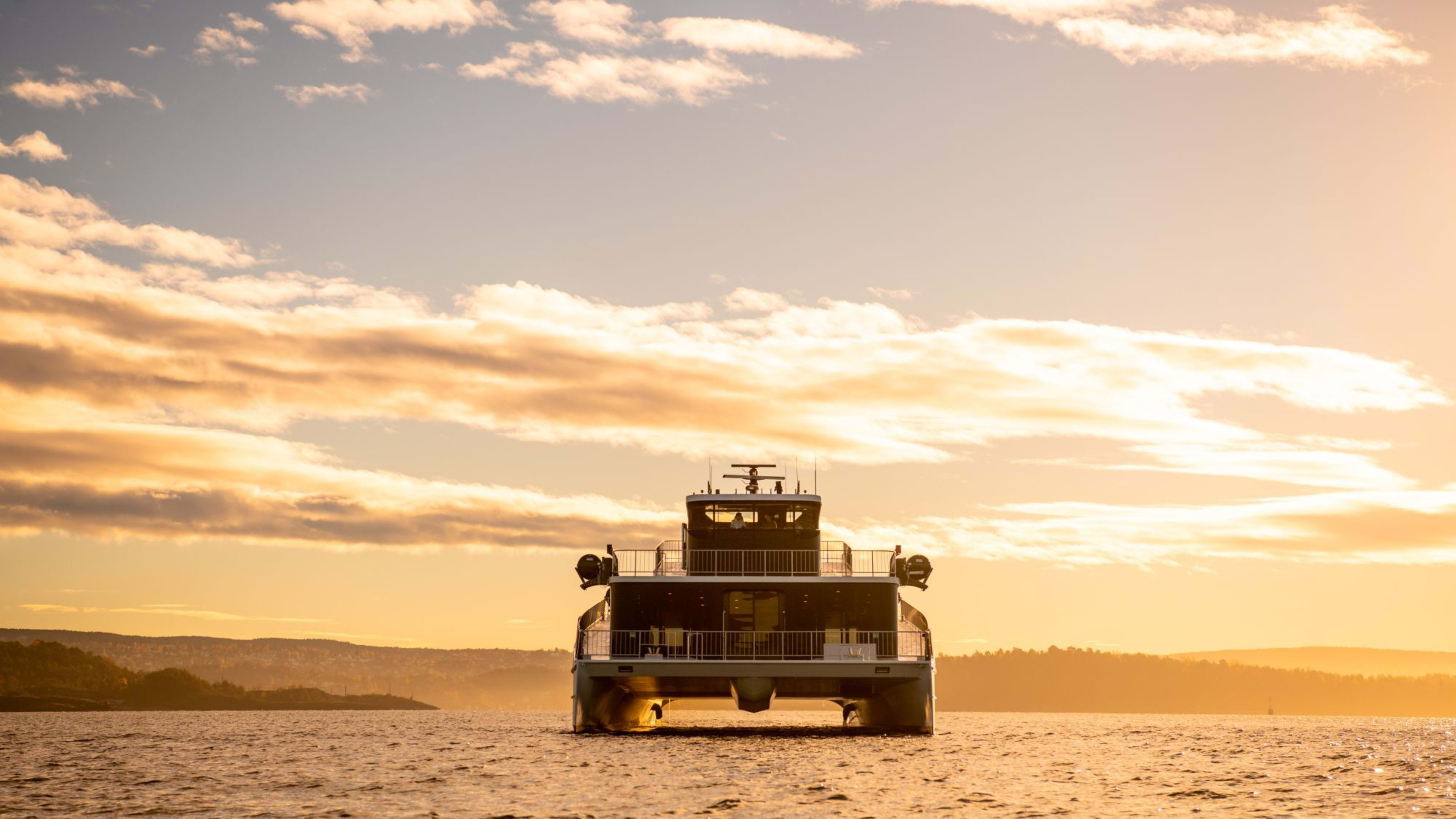 Ferje bakfra ved solnedgang med varme farger og svak kystlinje i horisonten.