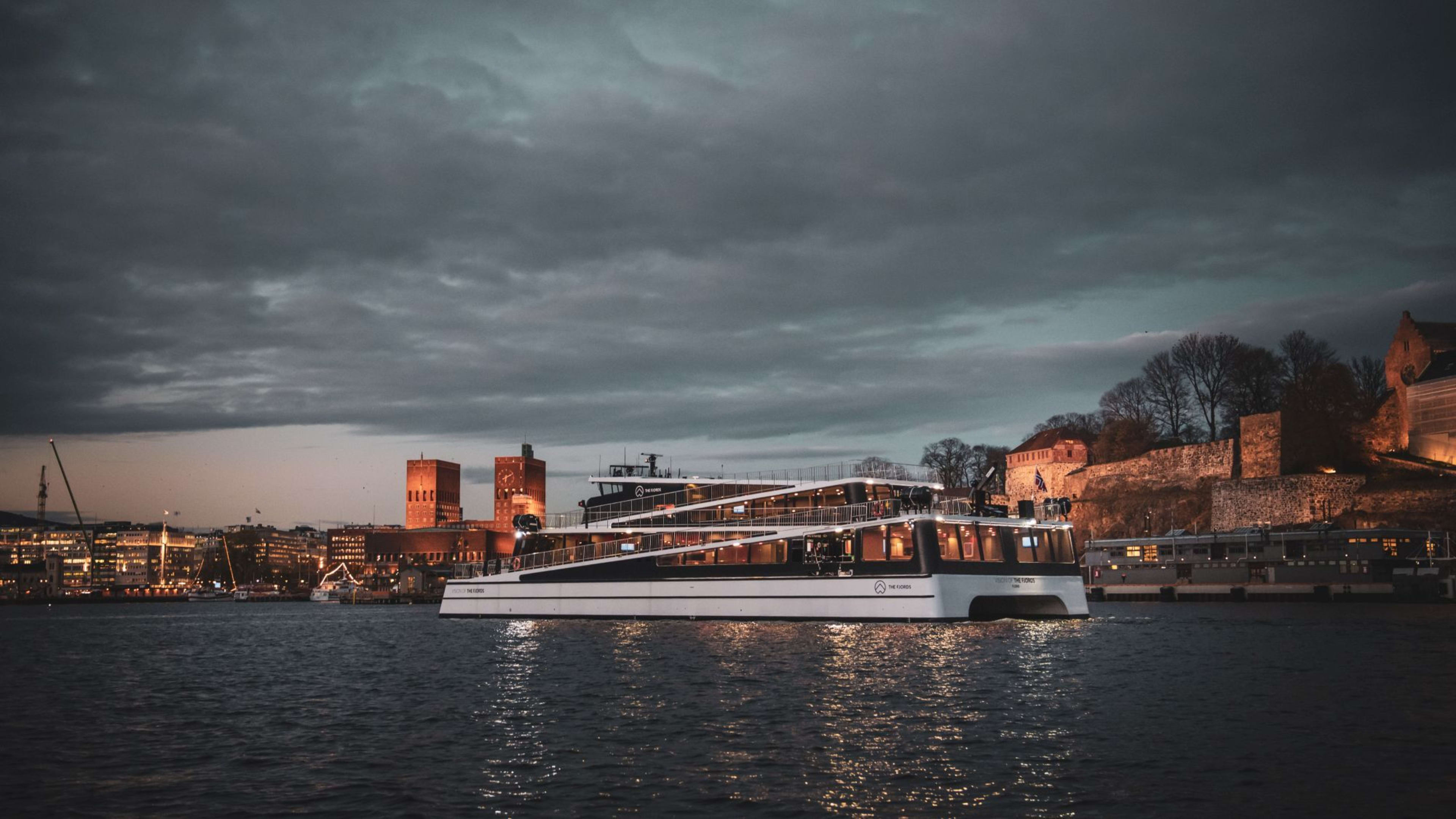 Nattferje passerer Akershus festning, med opplyst by og speilbilder i vannet.