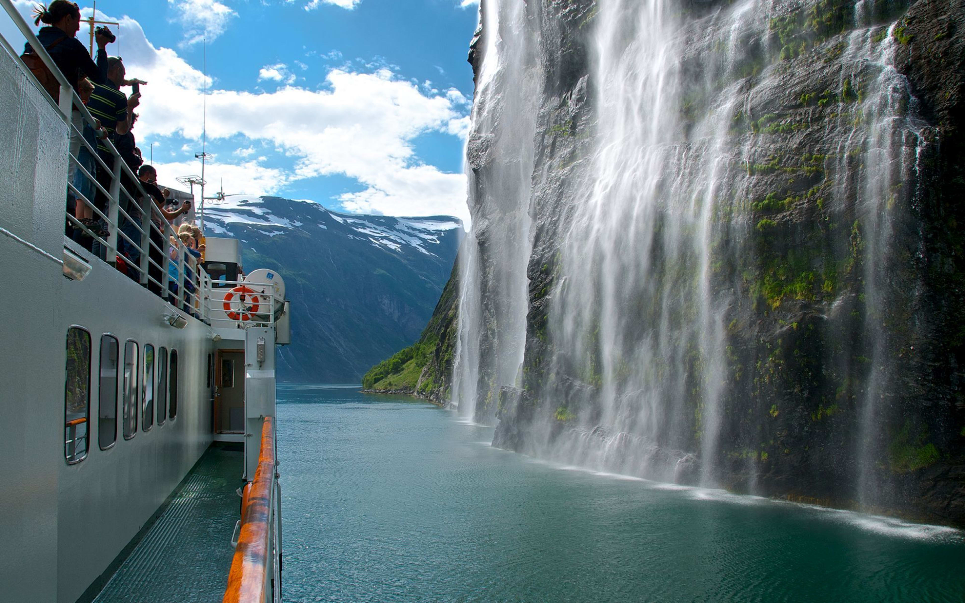 Tourists photograph waterfall from boat on fjord.