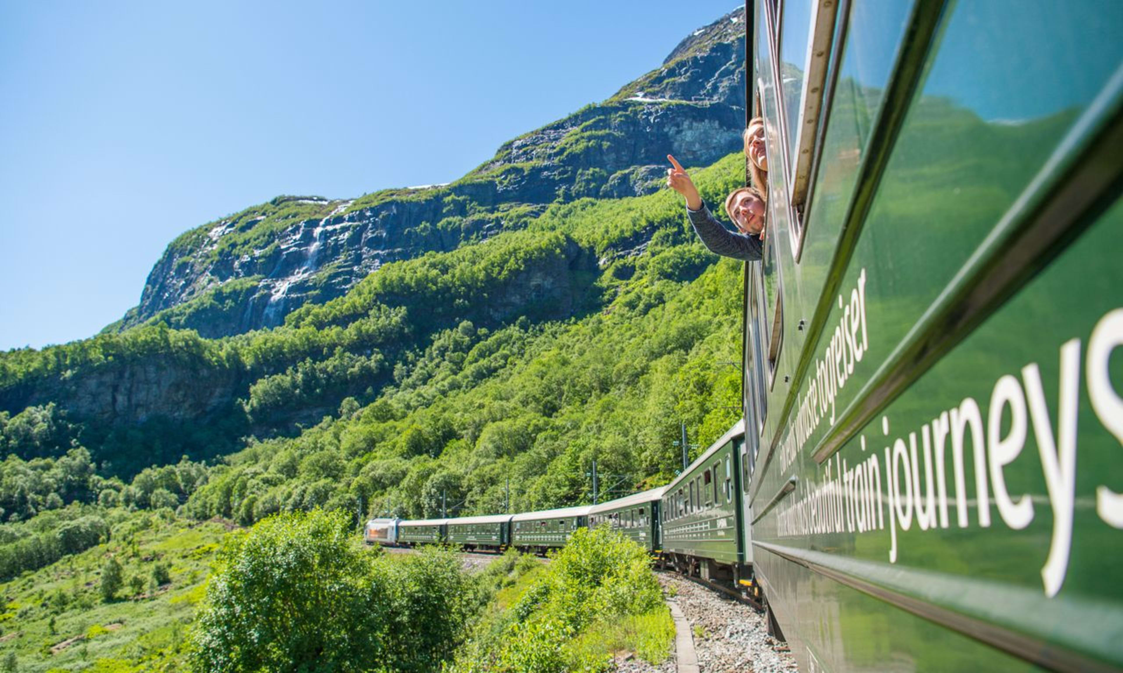 Passagiere lächeln aus Fenstern eines grünen Zuges, der durch Berge windet.
