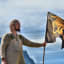 Man in Viking-style clothing holding black flag with Nordic artwork, dramatic mountain peaks in background and cloudy sky.