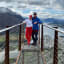 Par på utsiktsplattform med fjord og fjell i bakgrunnen under skyet himmel.