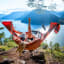 Person in orange hammock above fjord, legs up, sunny mountain view.