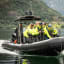 Tourists in yellow ride a boat through fjord with green hills and village.