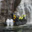 Ein schwarzes Festrumpfschlauchboot liegt in der Nähe eines Wasserfalls im Lysefjord. Die Passagiere heben aufgeregt die Arme.