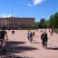 Cyclists and pedestrians approach a yellow palace in a sunny park.