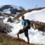 Smilende turgåer i blå skjorte og ryggsekk står ved skummende fjellbekk, med røft landskap og smeltende snø.