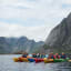 Group of kayakers in colorful kayaks on calm water, with jagged mountain peaks rising and cloudy skies in the background.