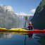 Smiling kayaker paddling a yellow and red kayak on calm water, with cliffs and lush hills surrounding a narrow fjord.