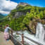 Girl leans on railing watching waterfall in forested cliffs and mountains under a vibrant blue sky.