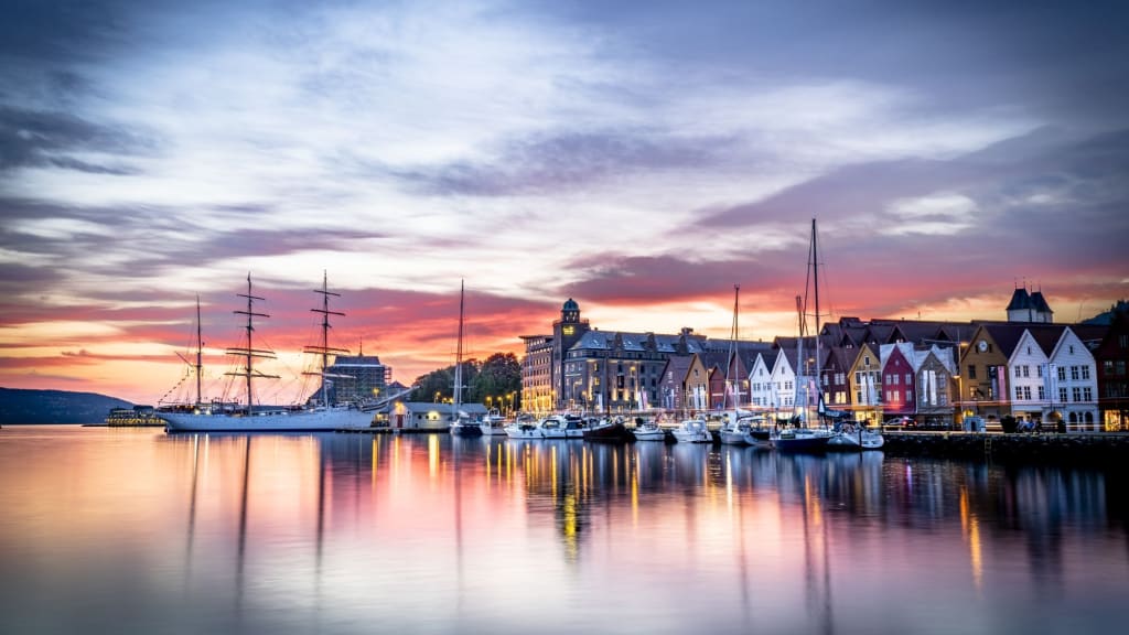 Sunset harbor scene with colorful wooden buildings, sailboats docked, and their reflections in calm water under a dramatic sky.