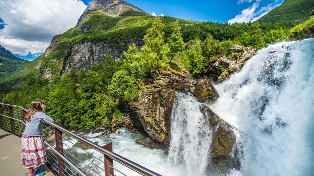 Jente lener seg mot rekkverk og ser på fossen i skog og fjell under sterk blå himmel.