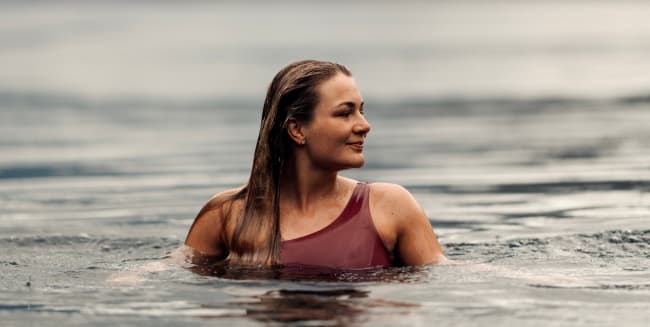 Woman swimming calmly in fjord, surrounded by misty mountains and lush green hills.