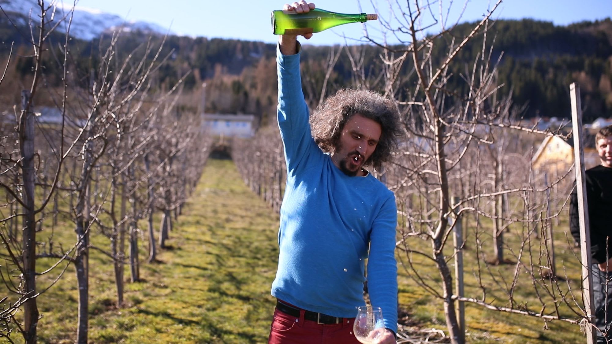 Man playfully pouring cider from height in orchard with apple trees and mountains behind, another smiling nearby.