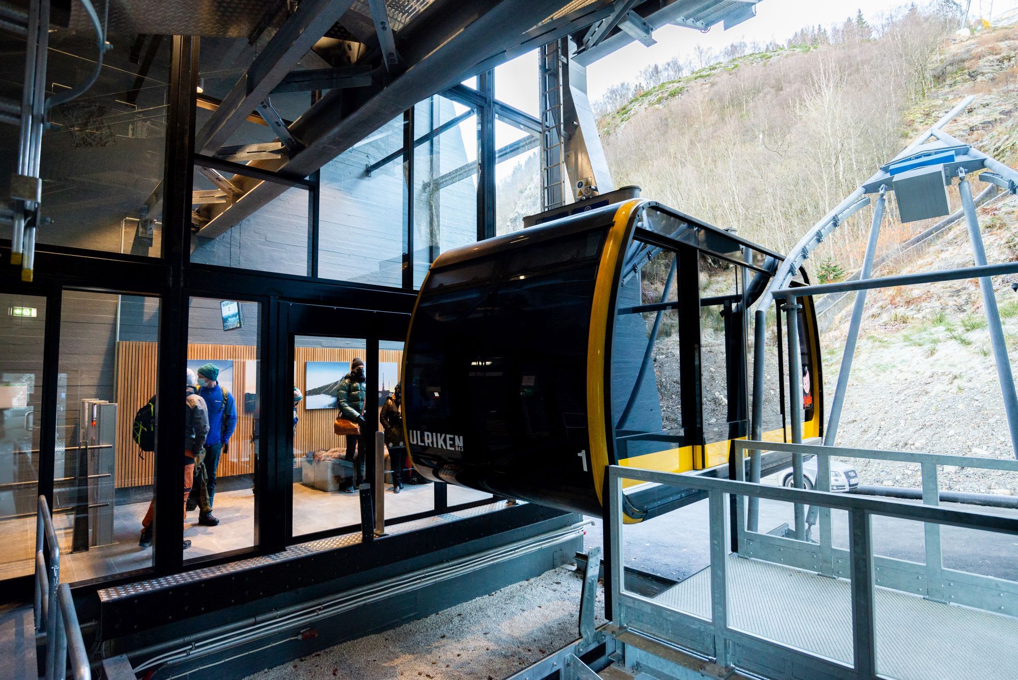 Moderne kabelvogn med glassvinduer på stasjon, passasjerer synlige og trær på fjellside i bakgrunnen.