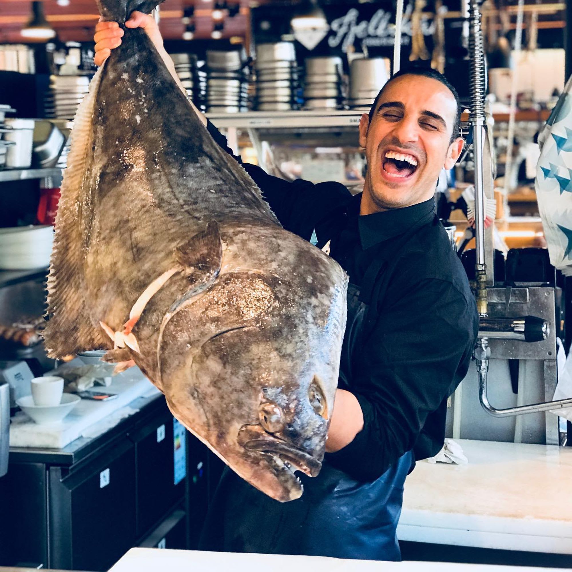 Ein fröhlicher Koch hält einen großen Heilbutt, lächelnd in einem lebhaften Markt mit Küchengeräten im Hintergrund.