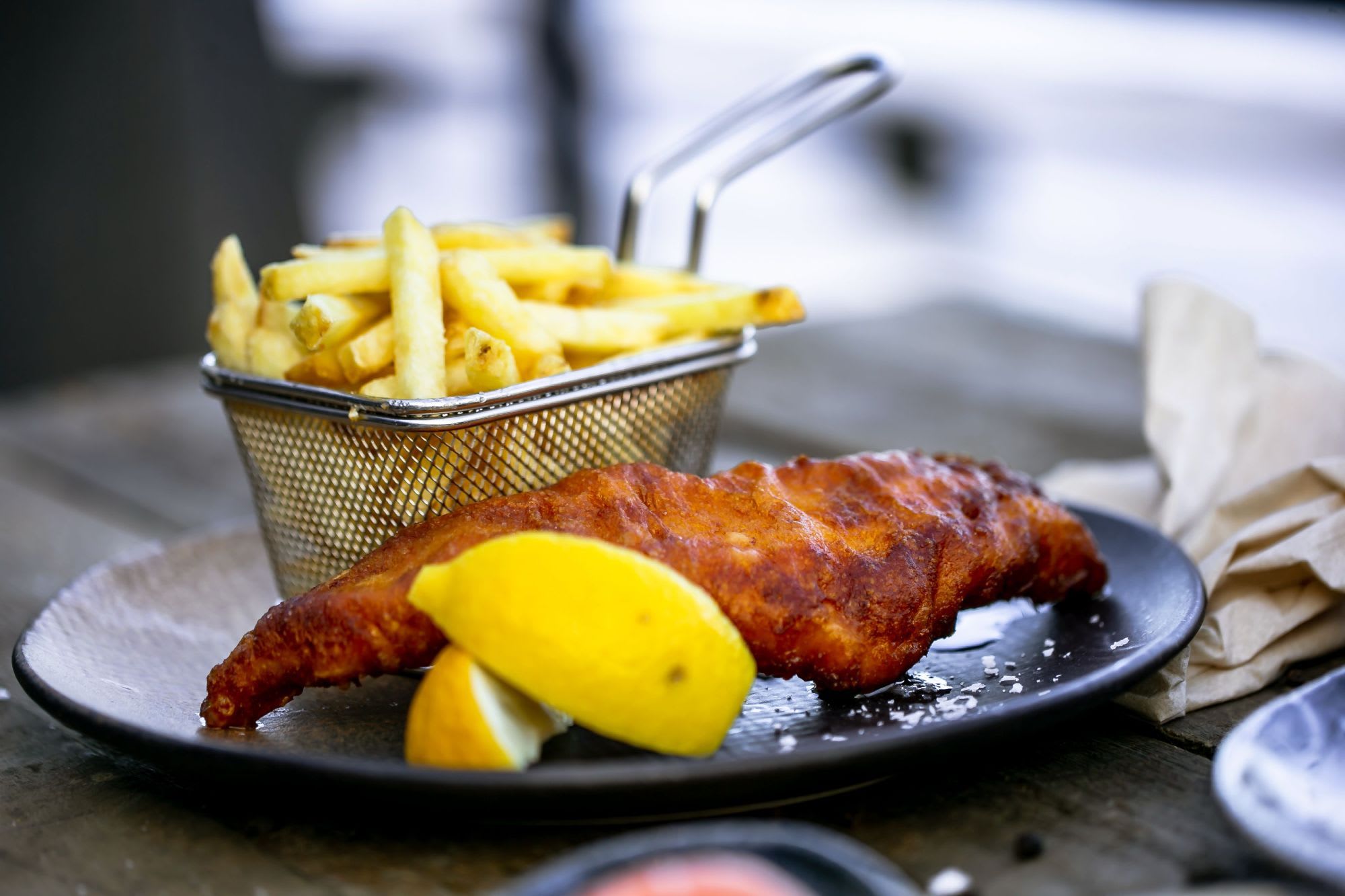 En klassisk fish and chips rett servert på en svart keramikkplate med sitronbåter, pommes frites og havsalt.