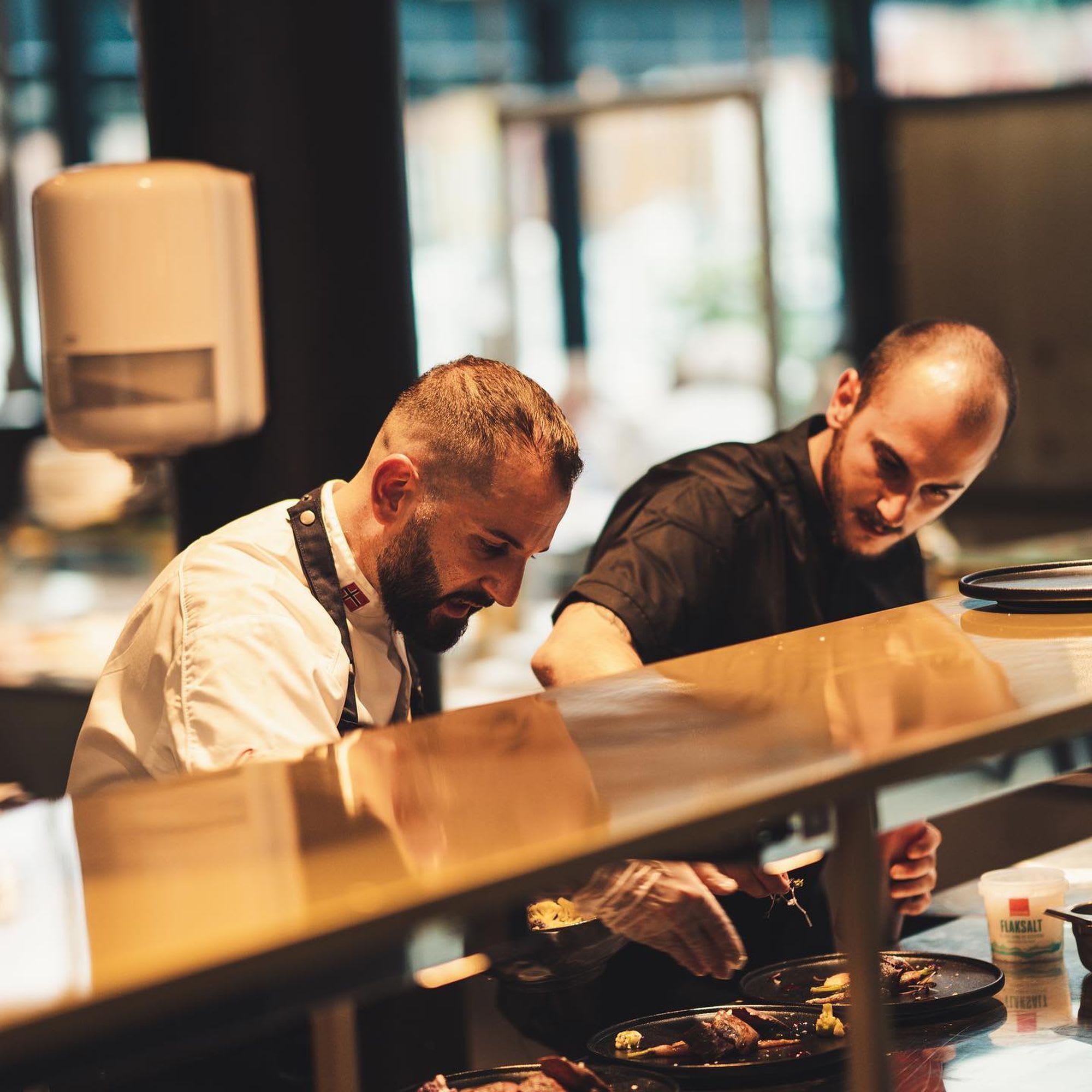 To kokker jobber i et moderne kjøkken, anretter retter og legger til finishing touches, med varmt lys.