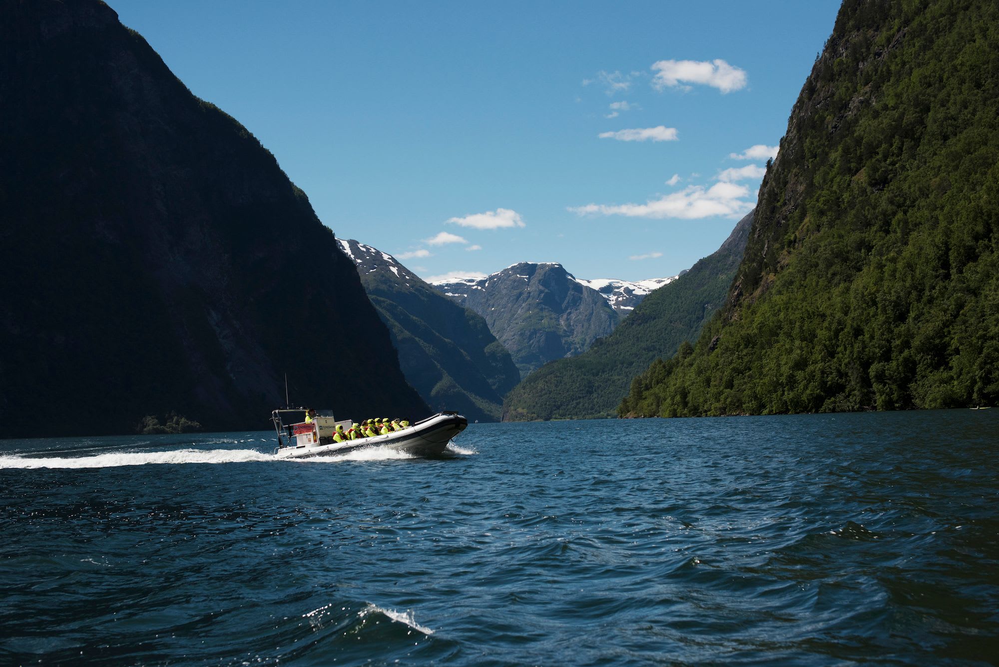 Hurtig oppblåsbar båt med passasjerer i redningsvester, seiler gjennom fjord med snødekte fjell i bakgrunnen.