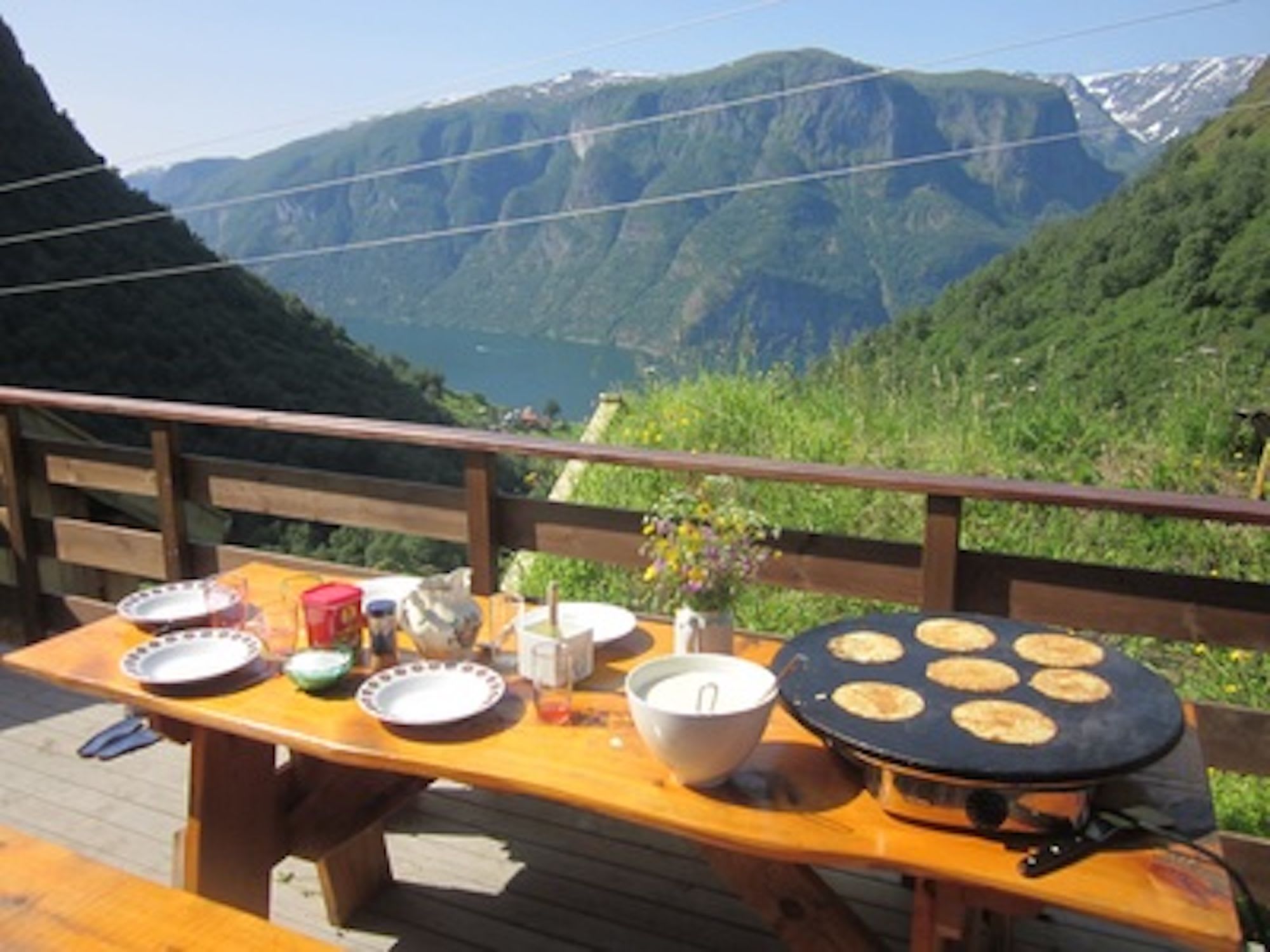 Utendørs rustikk bord med tallerkener, krydder, rørebolle og pannekaker som stekes, med fjellutsikt.