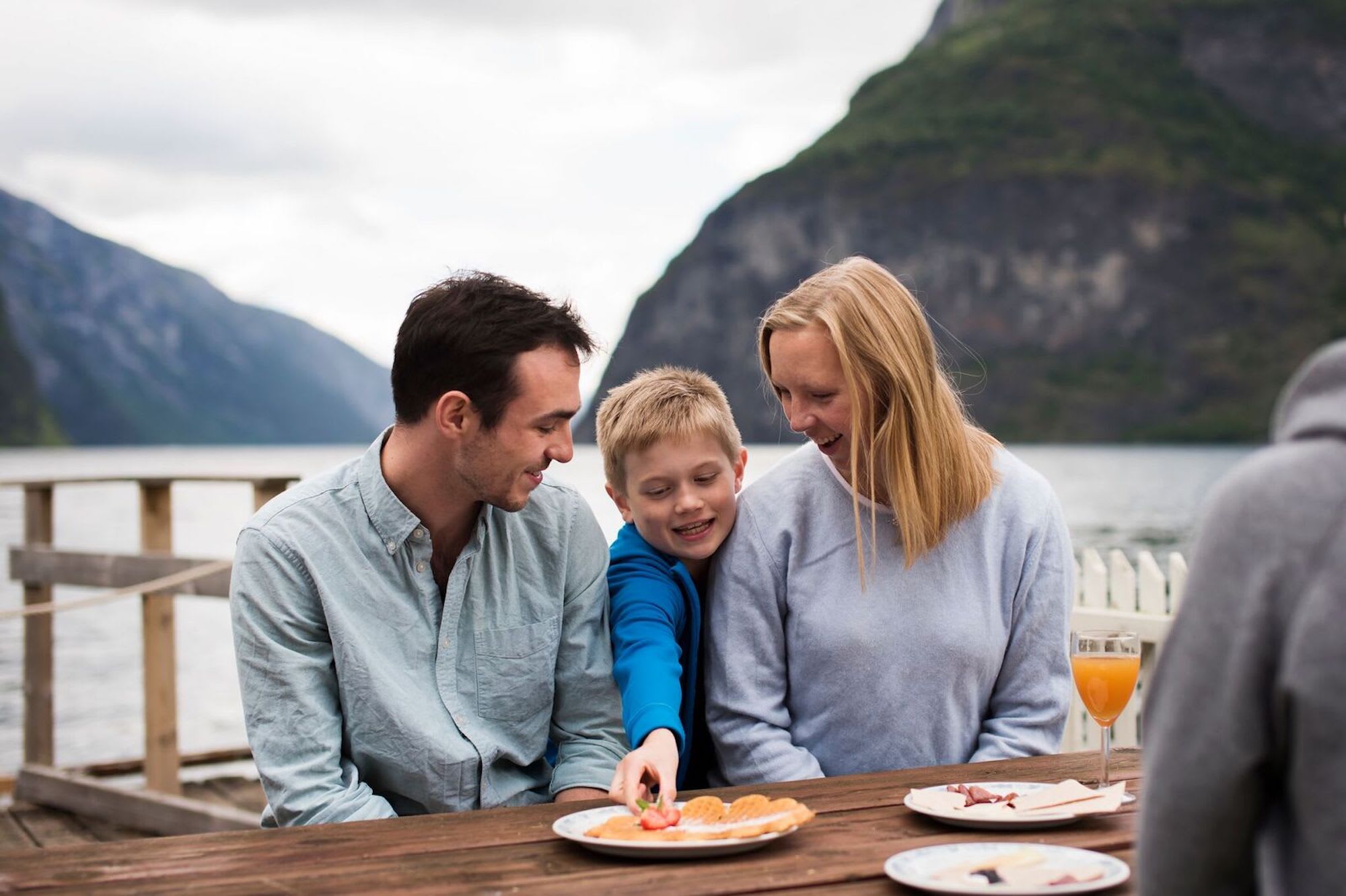 Familie von drei genießt eine Mahlzeit an einem Fjord, während ein Junge nach einer Erdbeere greift und Erwachsene lachen.