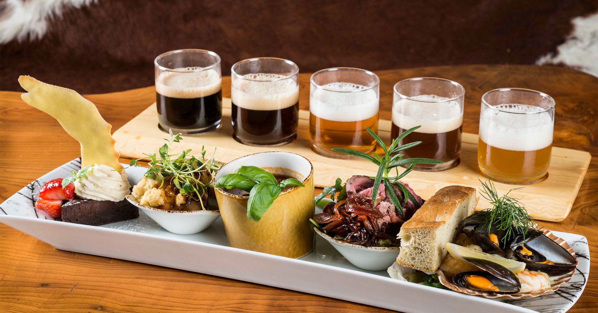 Gourmet platter with chocolate dessert, seafood, bread, and craft beers, served on a rustic wooden table with animal fur.