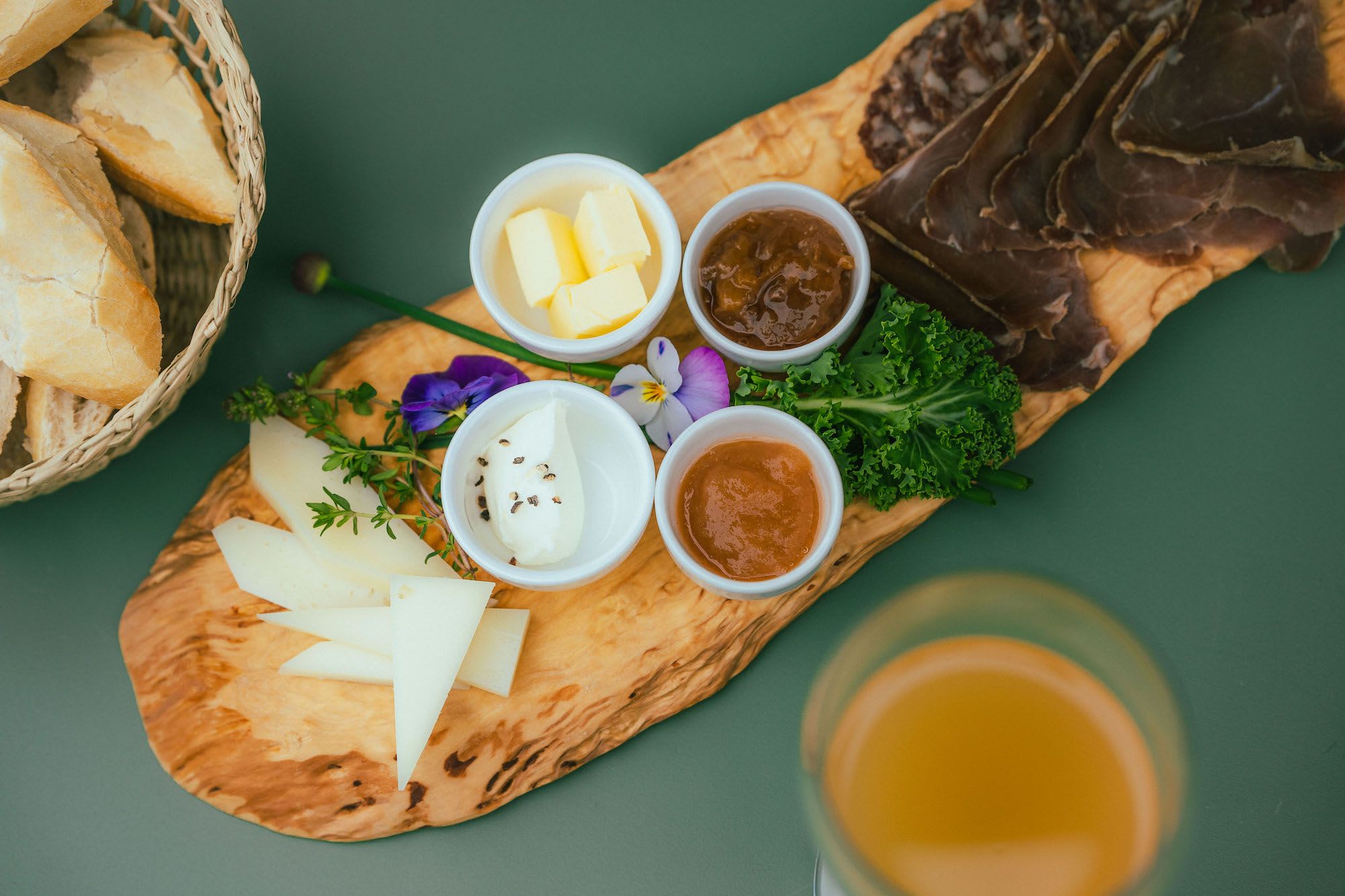 Trecharcuteriebrett med oster, spekemat, smør, pepperost, syltetøy, urter og brød, med cider i nærheten.