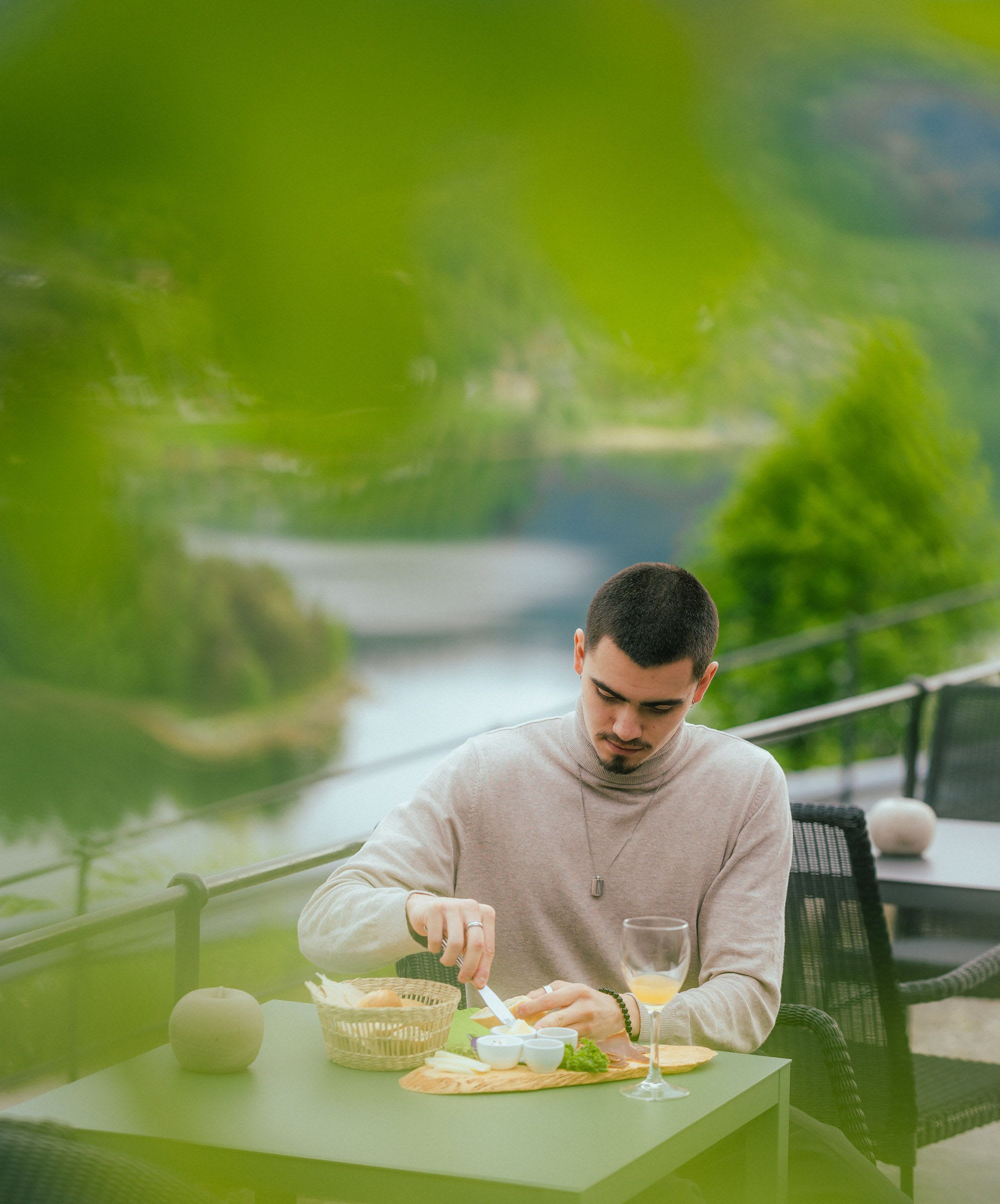 Mann smører smør på brød fra trebrett utendørs, med cider, sjøutsikt og grønn bakgrunn.