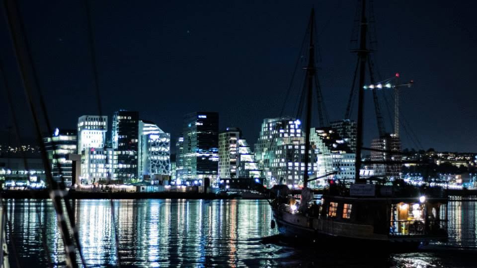Nattbilde av bylandskap med opplyste høyhus og seilbåt i forgrunnen mot en mørk himmel.