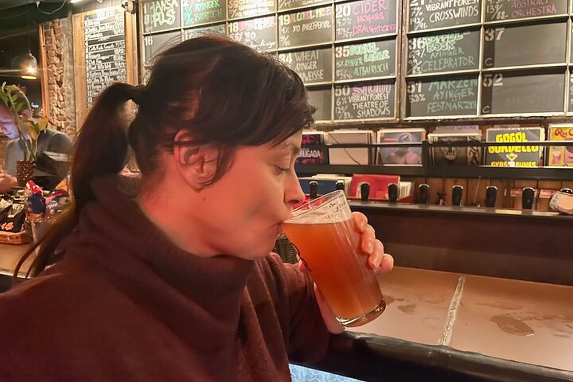 Kvinne som nyter amber håndverksøl på koselig bar, nyter smaken med lukkede øyne, varm belysning og rustikk innredning i bakgrunnen.