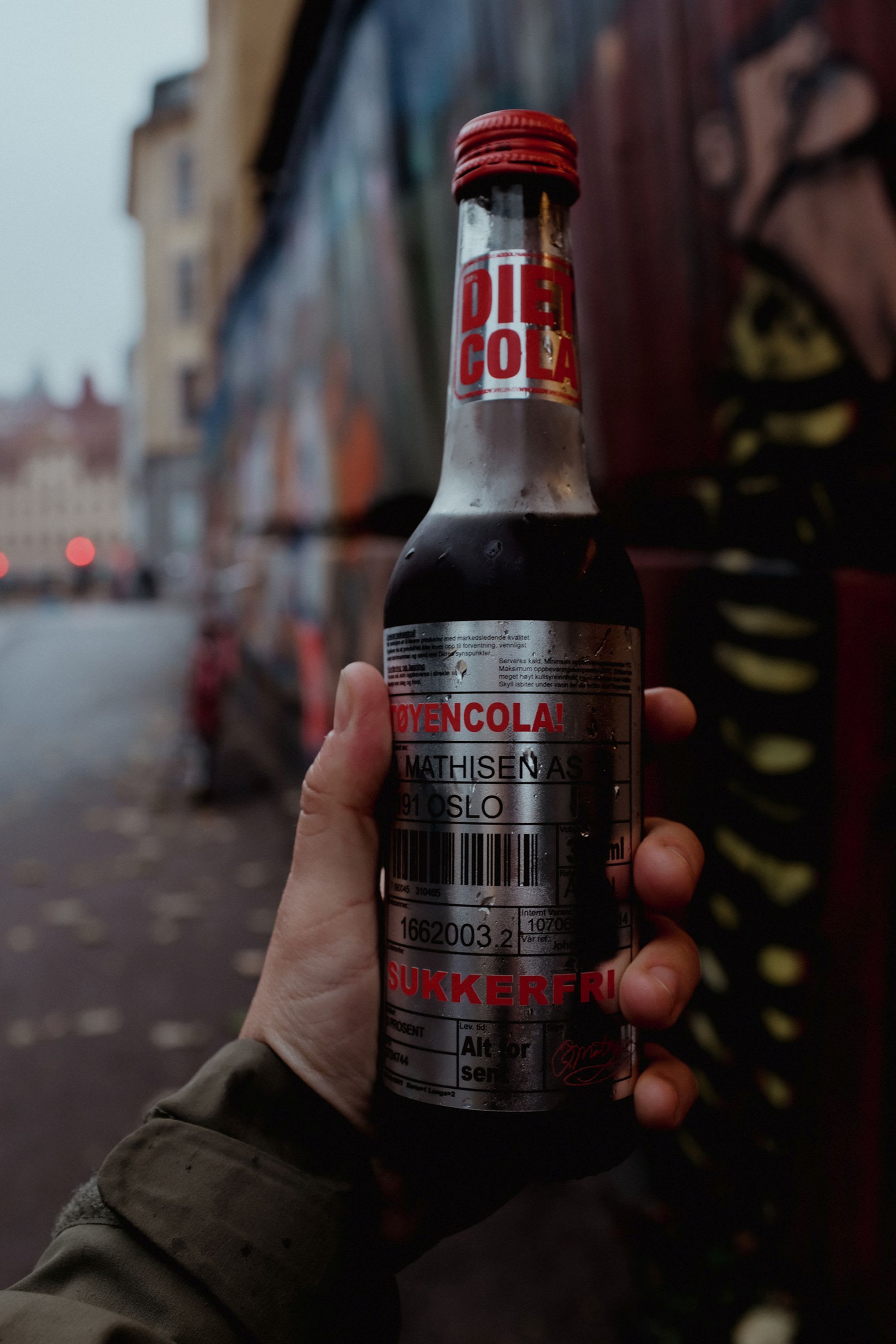 Nahaufnahme von Hand, die eine gekühlte "TøyenCola"-Flasche mit Silberetikett hält, vor einer Graffiti-bedeckten Wand im Hintergrund.