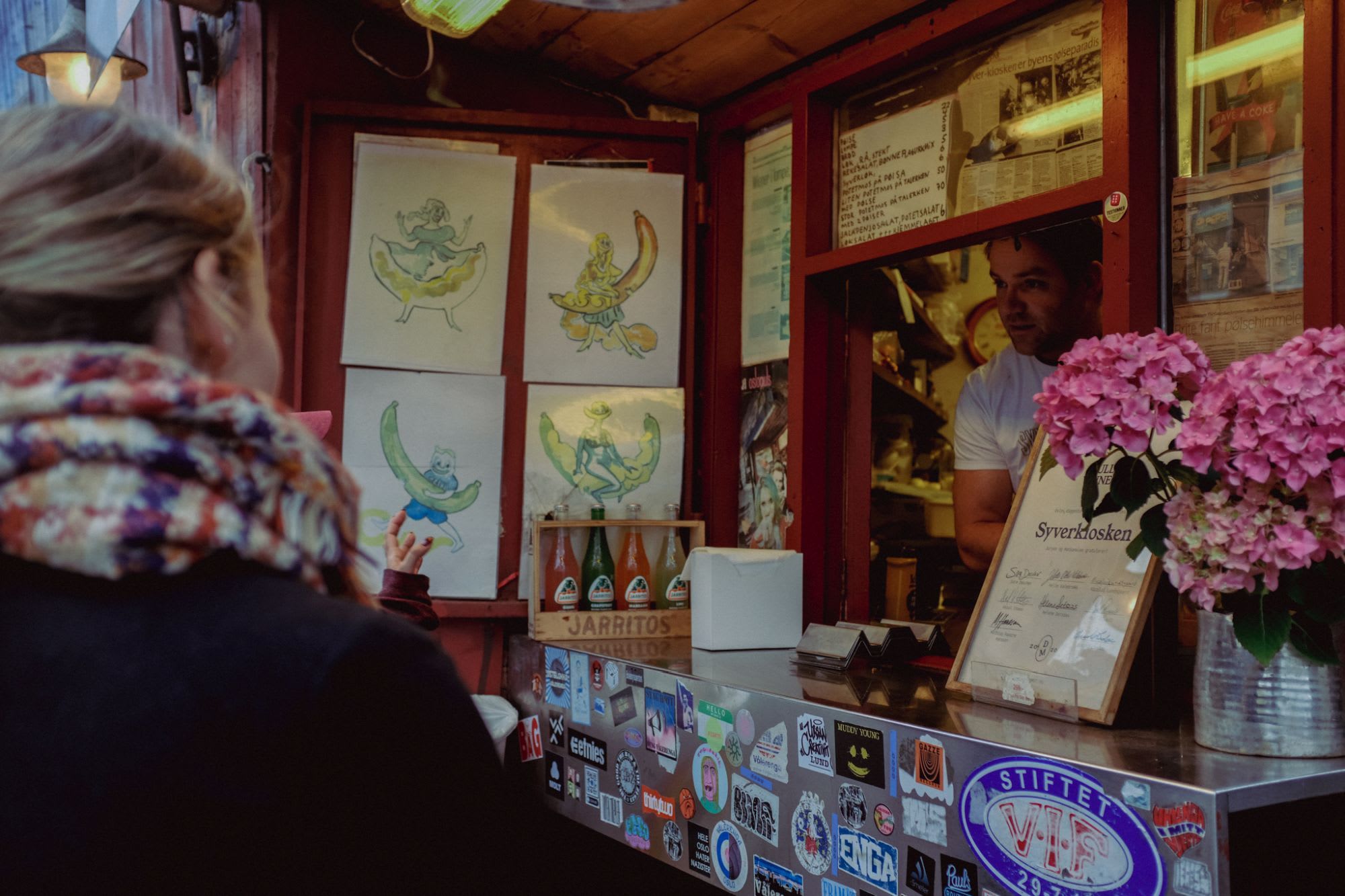 Skurrer Food-Kiosk mit buntem Bananen-Kunstwerk, freundlichem Verkäufer und eklektischer Dekoration mit Jarritos-Flaschen und rosa Blumen.