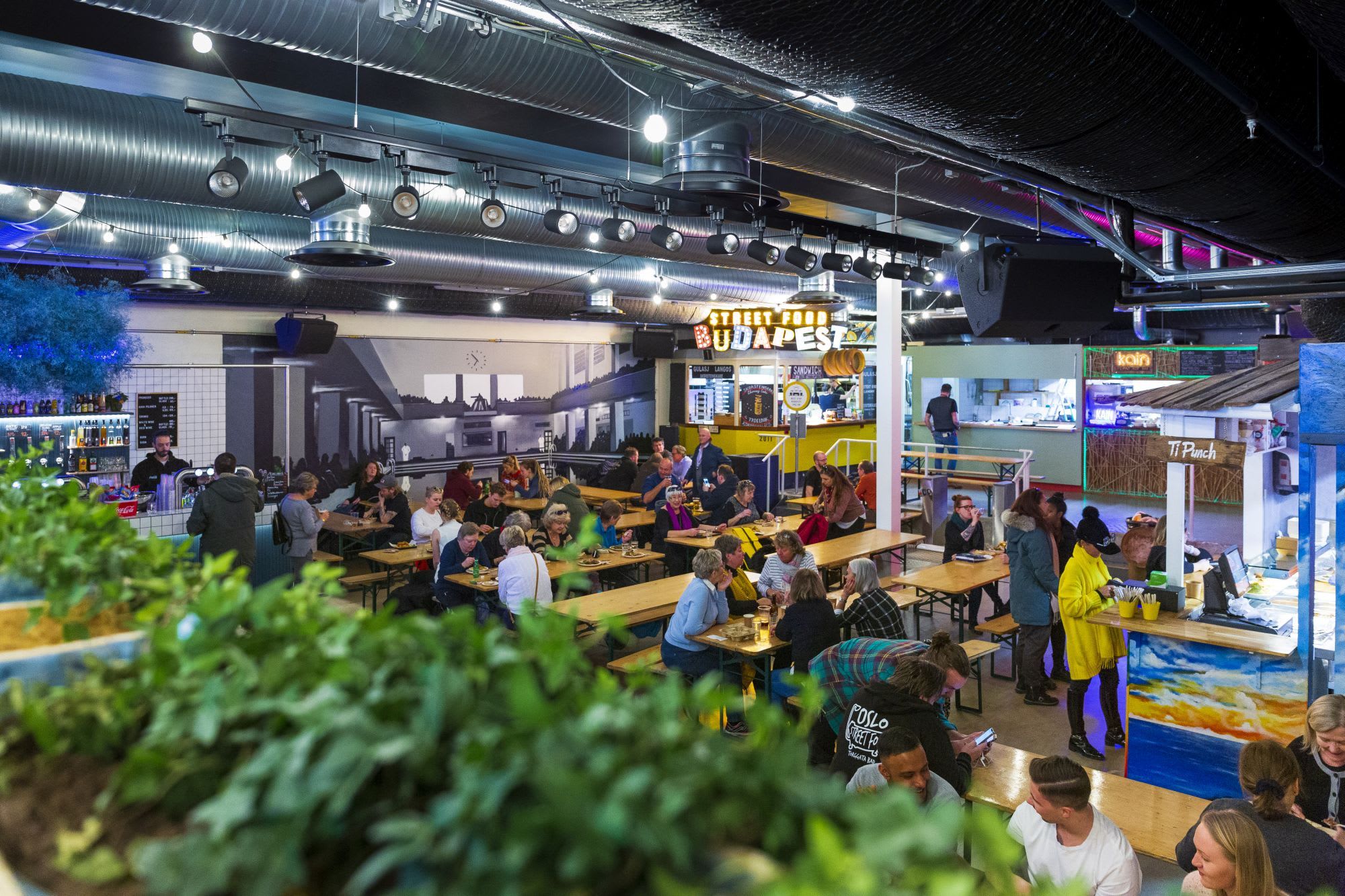 A vibrant street food market with colorful booths, tiki-themed bars, and communal seating for customers.