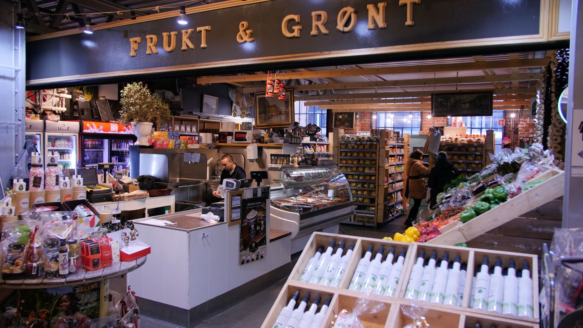 A vibrant indoor market stall with fresh produce, oils, and packaged goods, in a rustic setting with exposed brick.