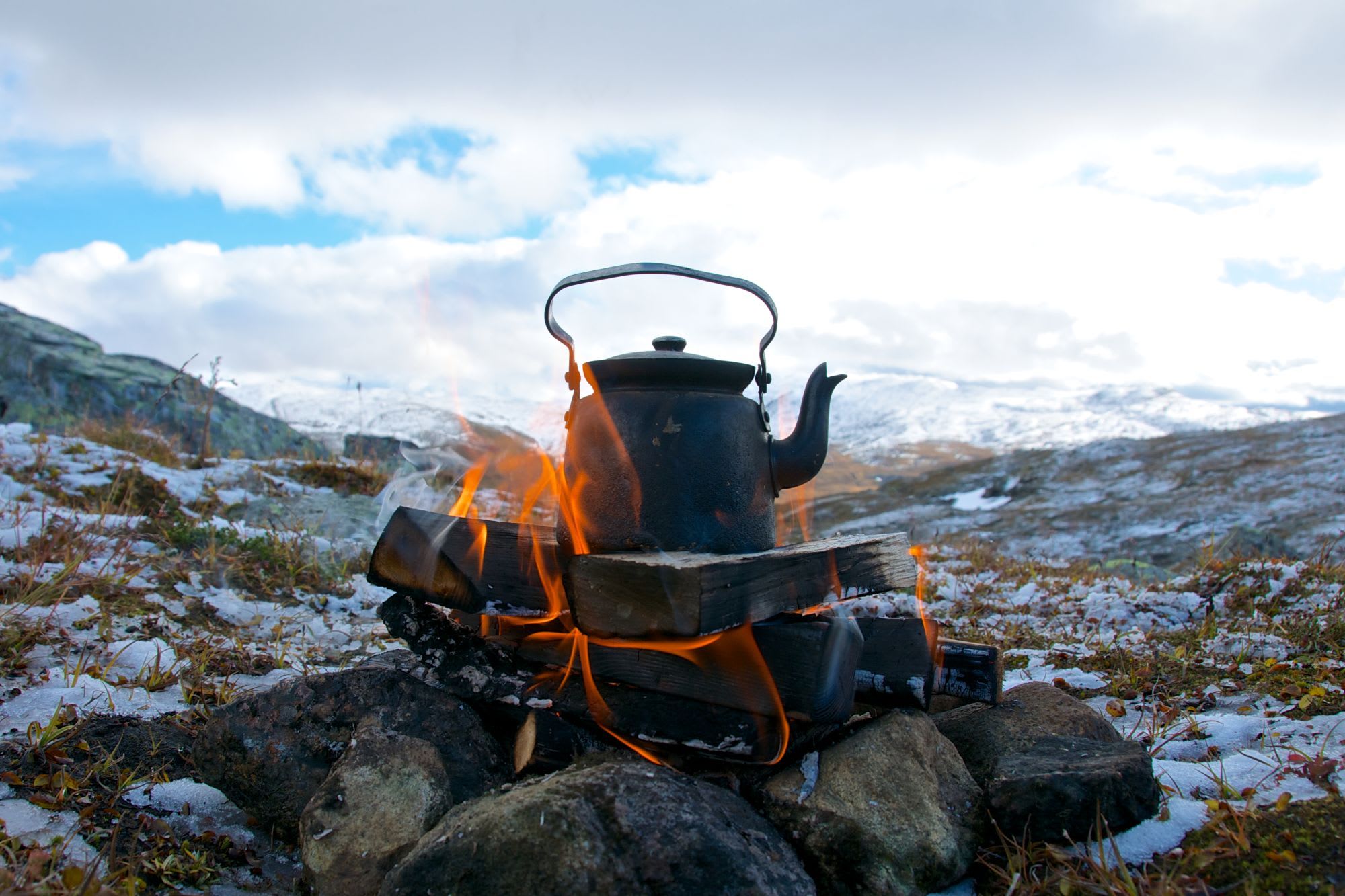 En svartkokegryte hviler på en vedfyrt ild med levende oransje flammer, omgitt av steiner og snødekt fjellbakgrunn.