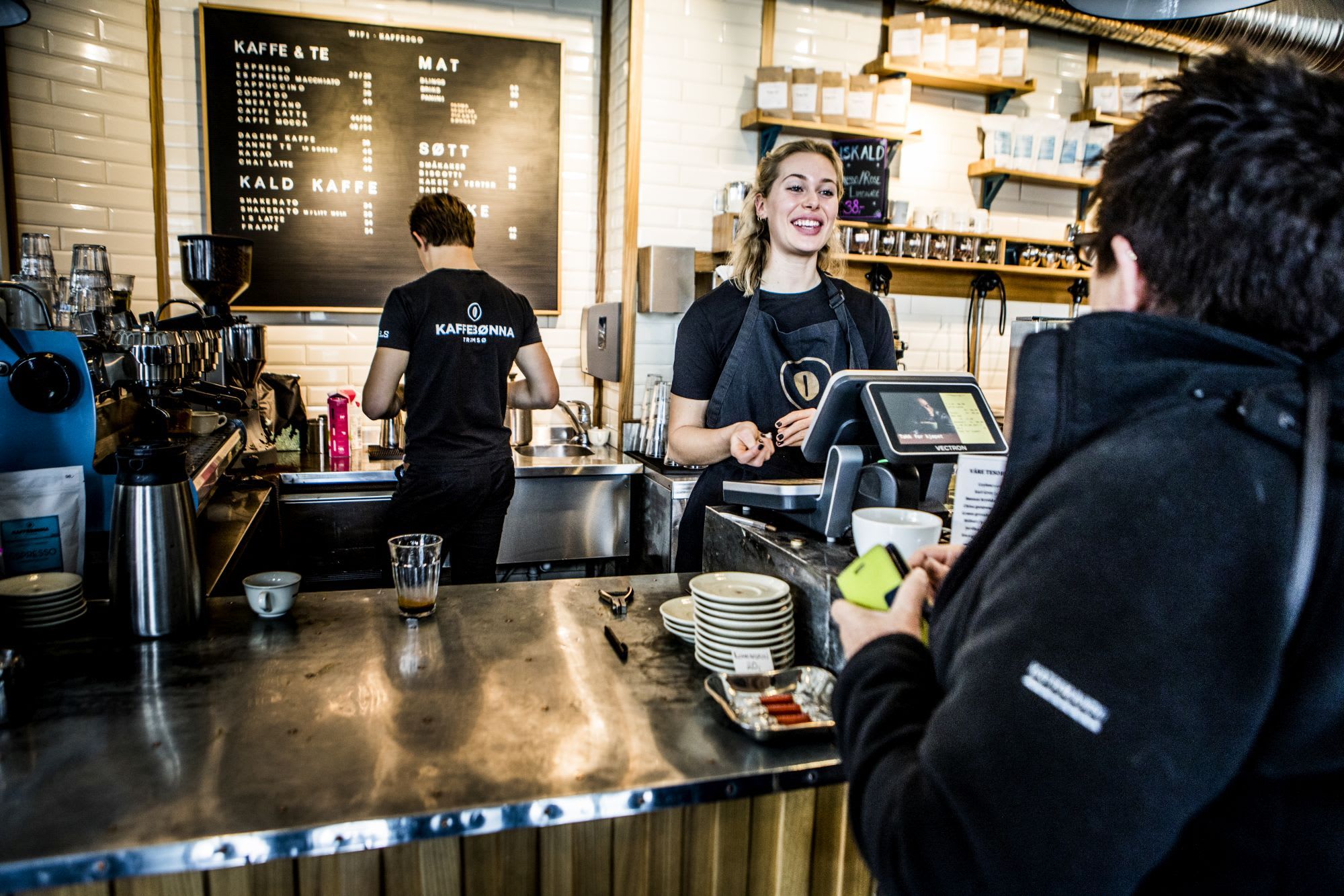 En smilende barista lager kaffe ved et polert disk med kaffekopper og en stor tavlemeny.