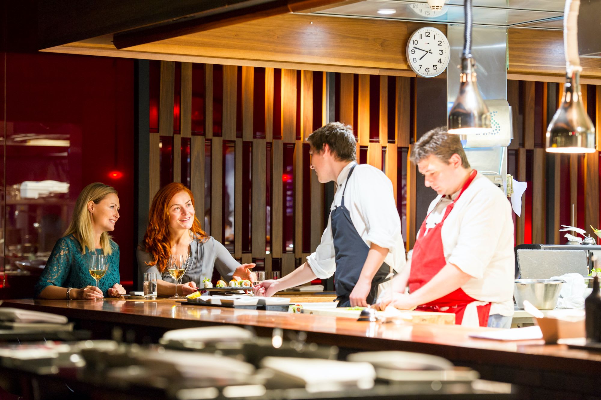 Frauen an der Restaurantbar, die mit dem Koch, der Sushi serviert, in einem modernen, warmen Küchenumfeld interagieren.