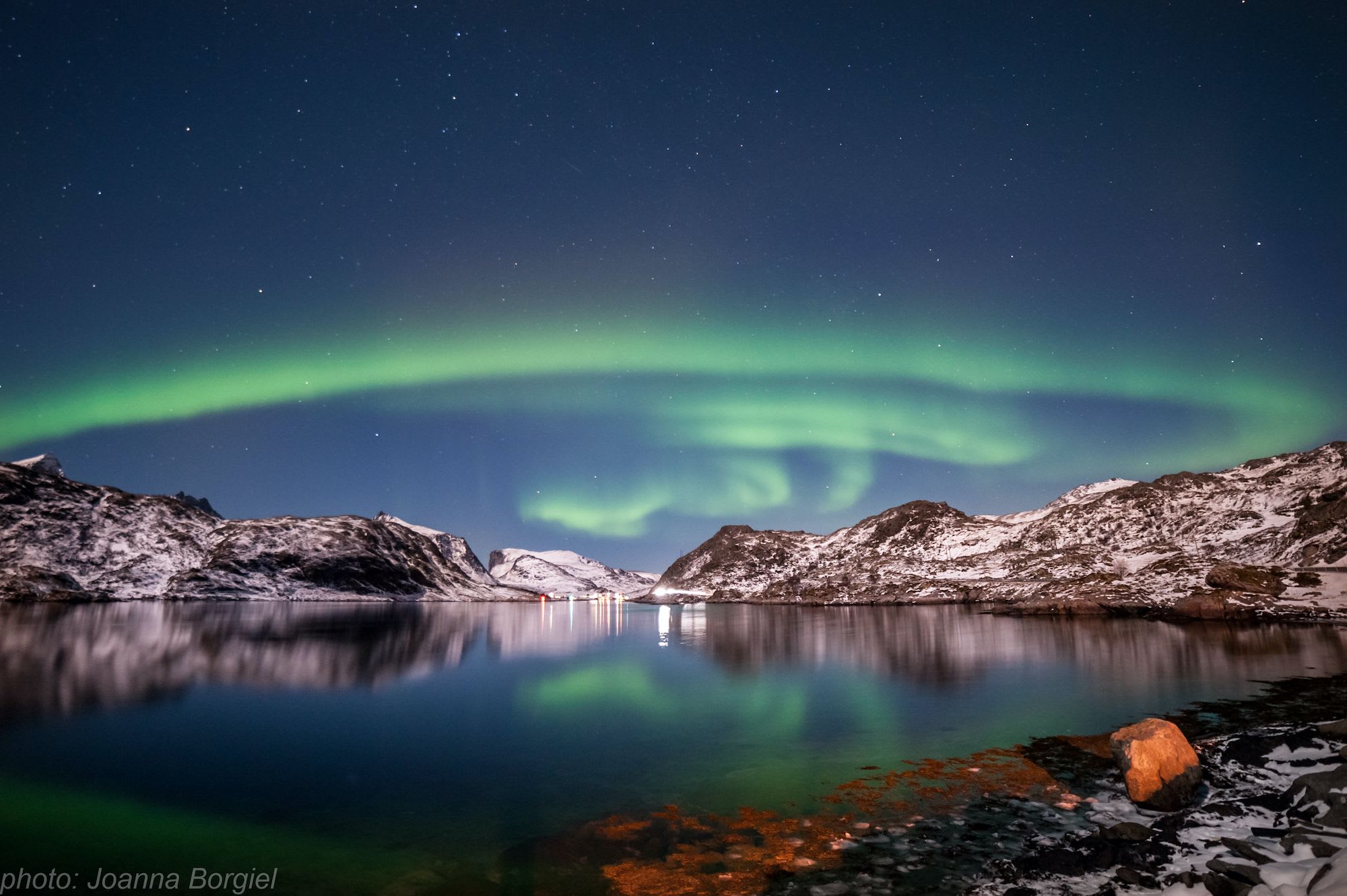 Fantastiske nordlys (Aurora Borealis) som lyser opp himmelen over snødekte fjell og stille vann.