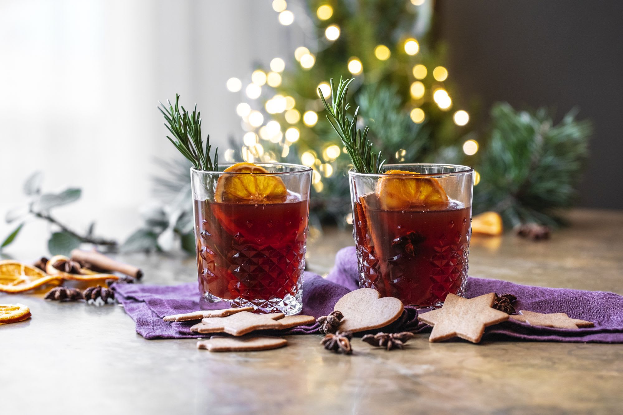 Krystallklare drikker med tørket appelsin og rosmarin, pepperkaker og krydder, satt mot blinkende julelys.